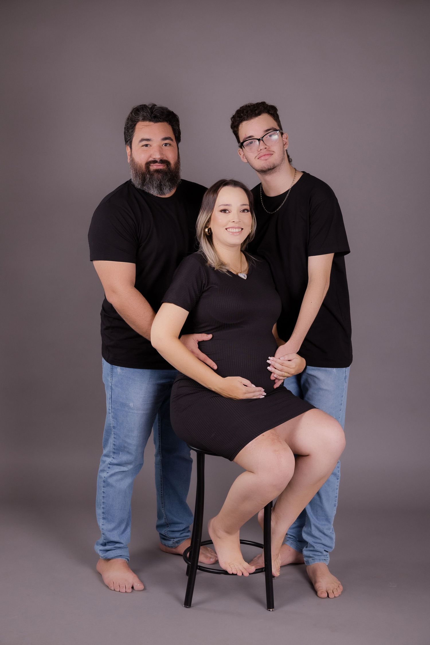 Three people in black outfits pose together for a maternity photo on a dark background.