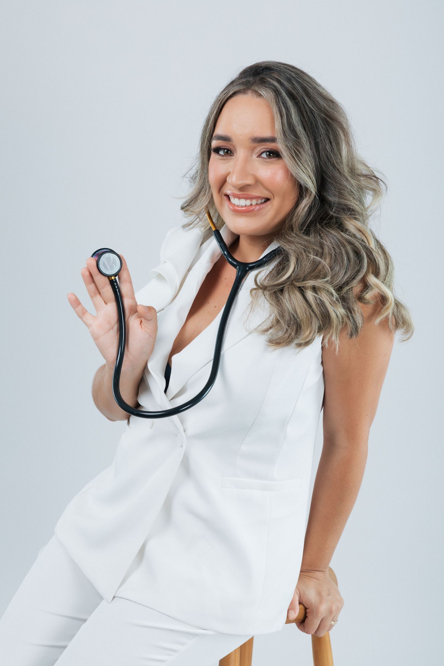A person in white medical attire holds a stethoscope while smiling against a light studio background.