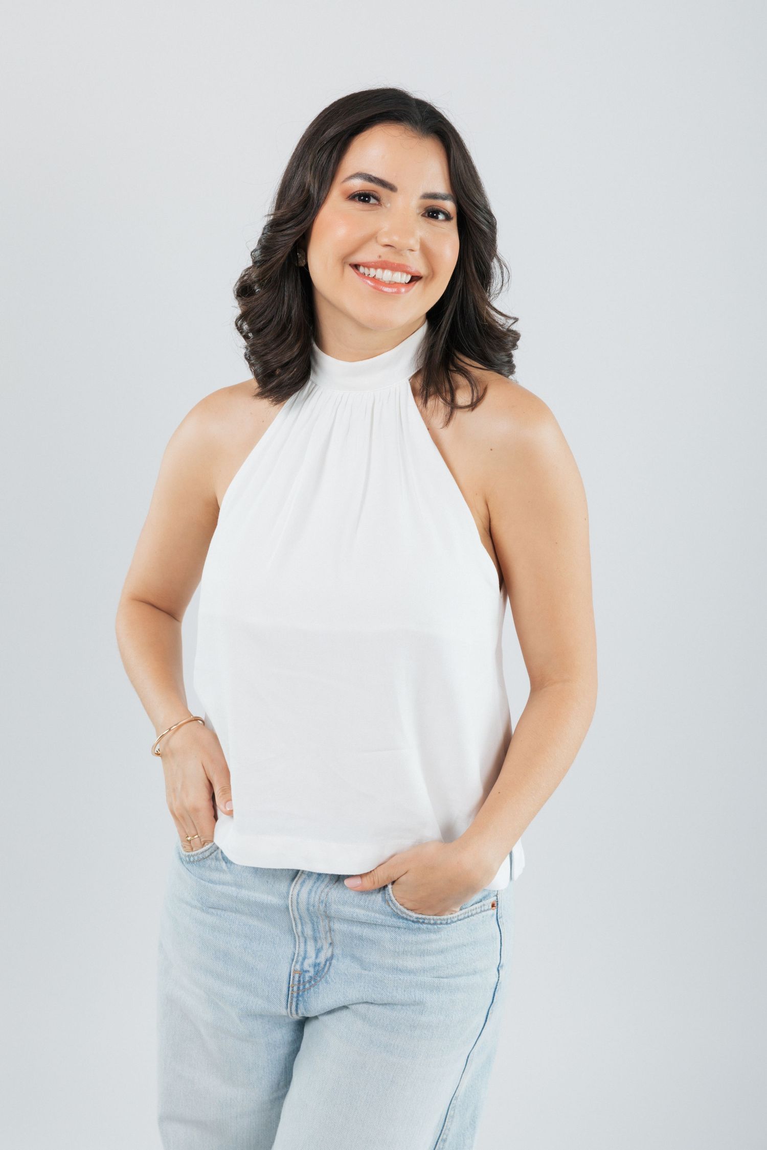 Professional model wearing white halter top and light denim jeans poses against light gray background.