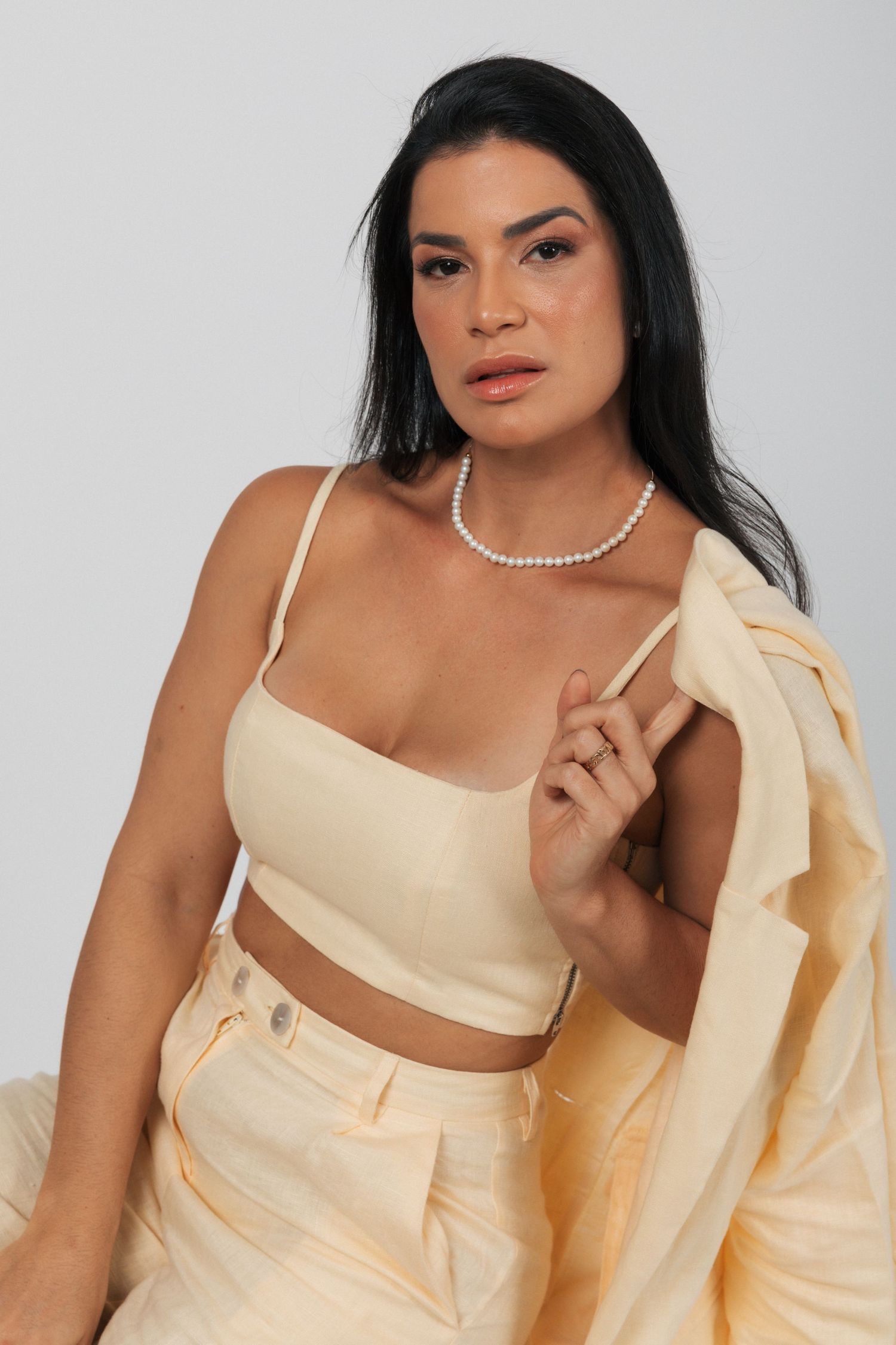 Fashion model poses in cream crop top and matching suit jacket with pearl necklace against white backdrop.