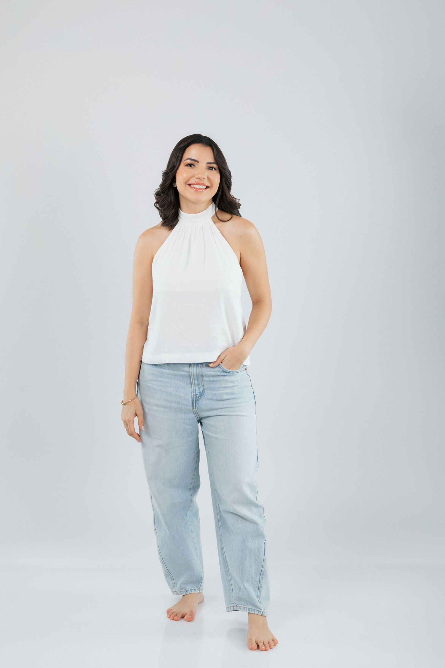 Person wearing white halter top and light blue jeans poses casually while standing barefoot against gray background.