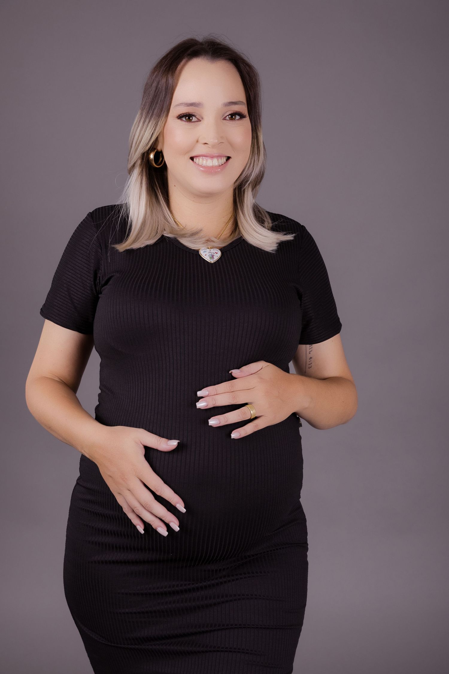 Pregnant woman wearing black dress poses against gray backdrop in maternity photoshoot.