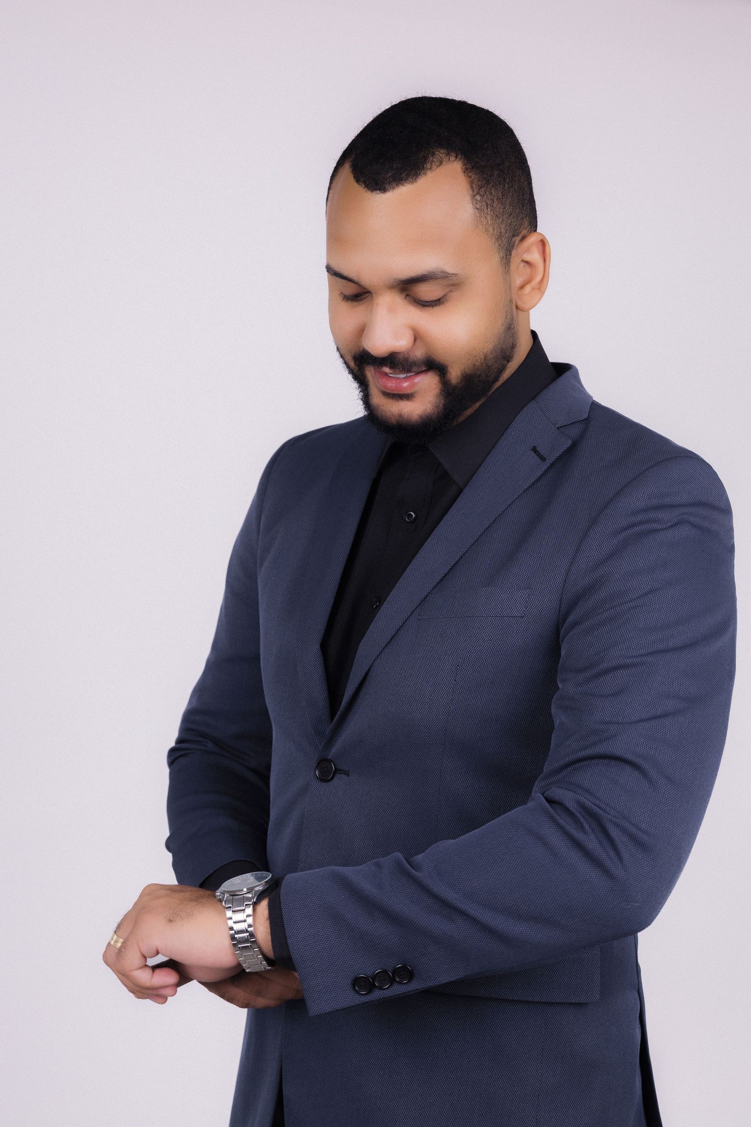 Professional wearing a navy suit and black turtleneck checks wristwatch while smiling against light background.