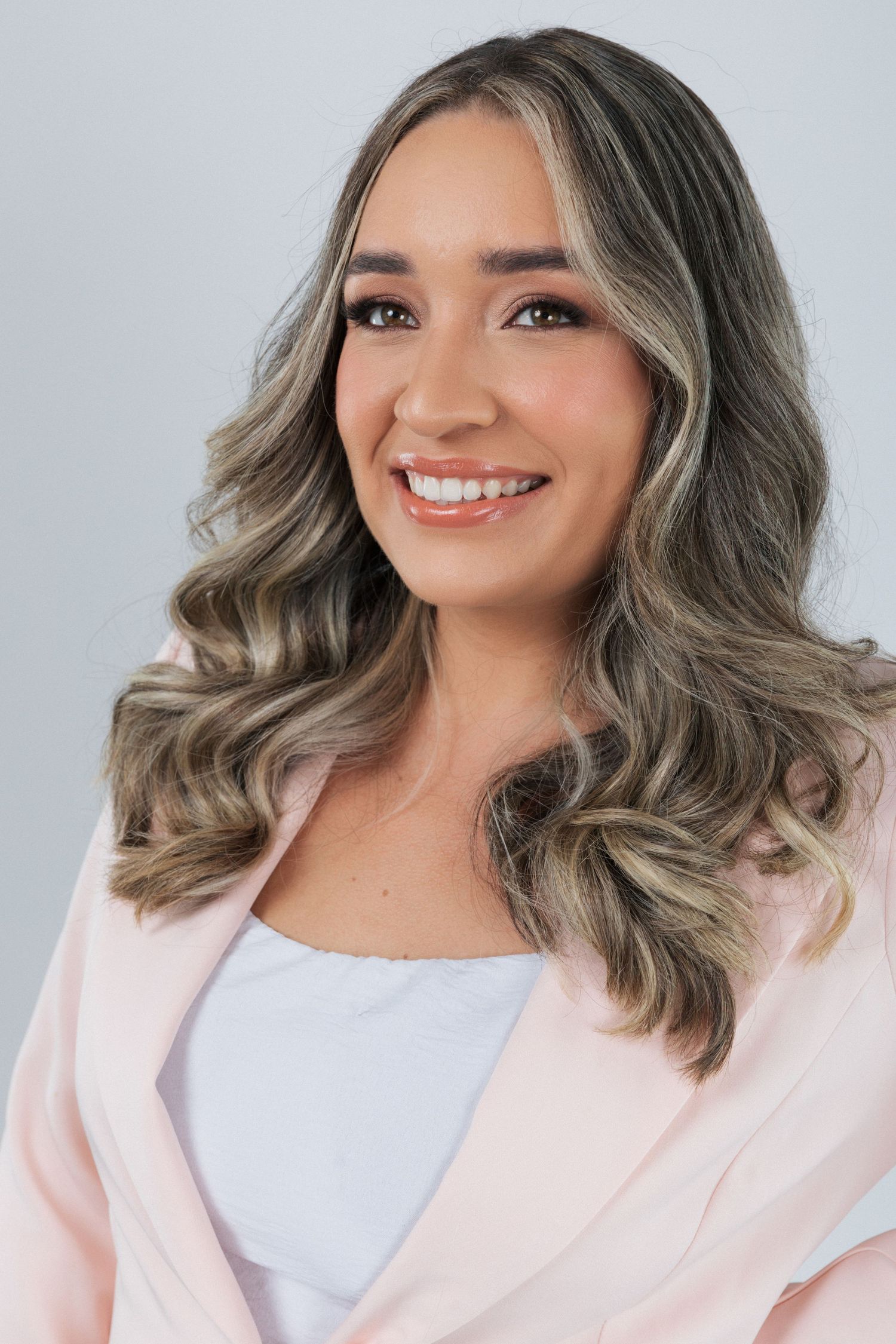 Professional portrait of person with wavy ash blonde hair wearing a light pink blazer and white top against gray backdrop.