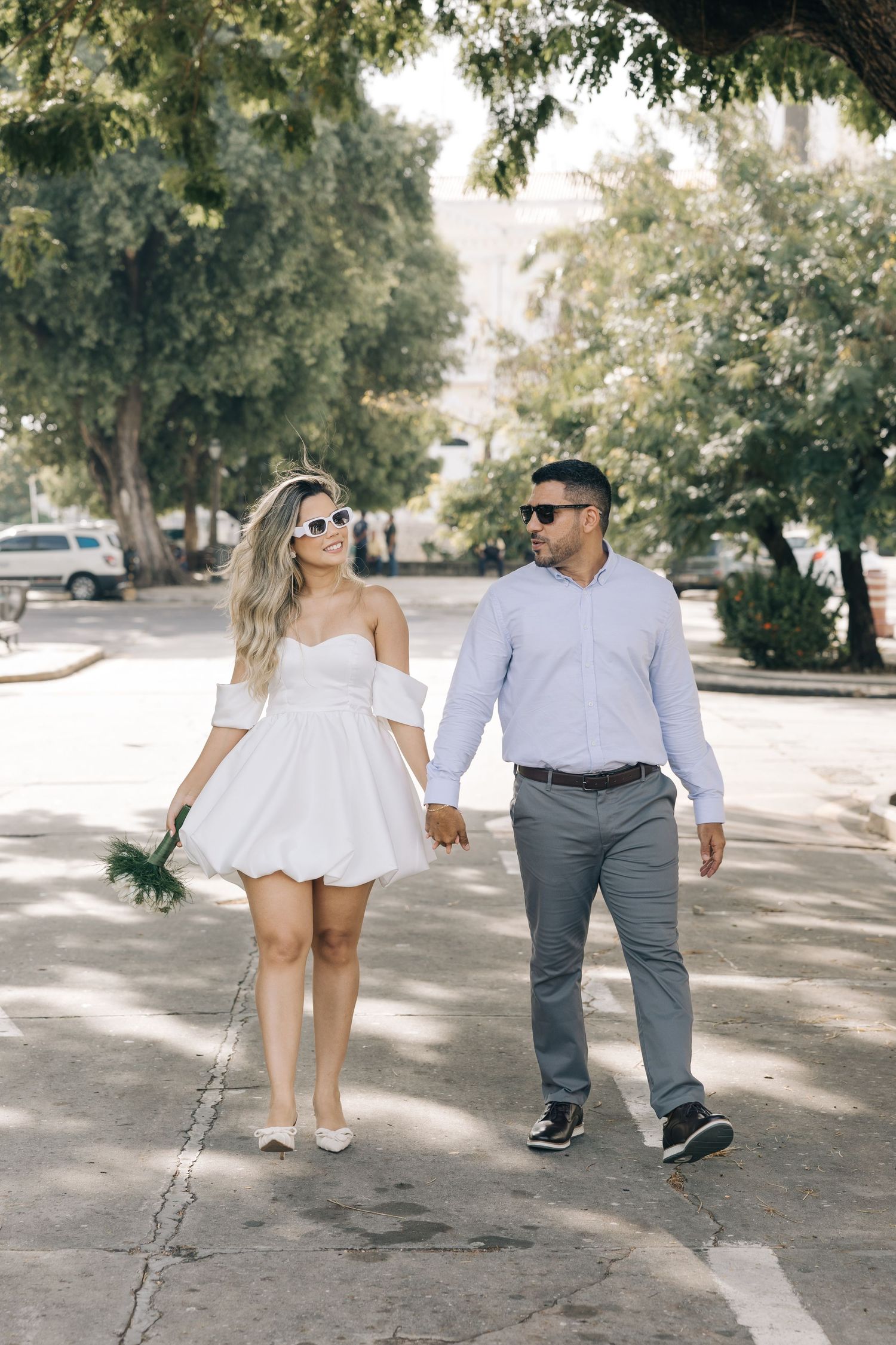 A couple walking together on a tree-lined street, with her in a white dress and him in a light blue shirt and gray pants.