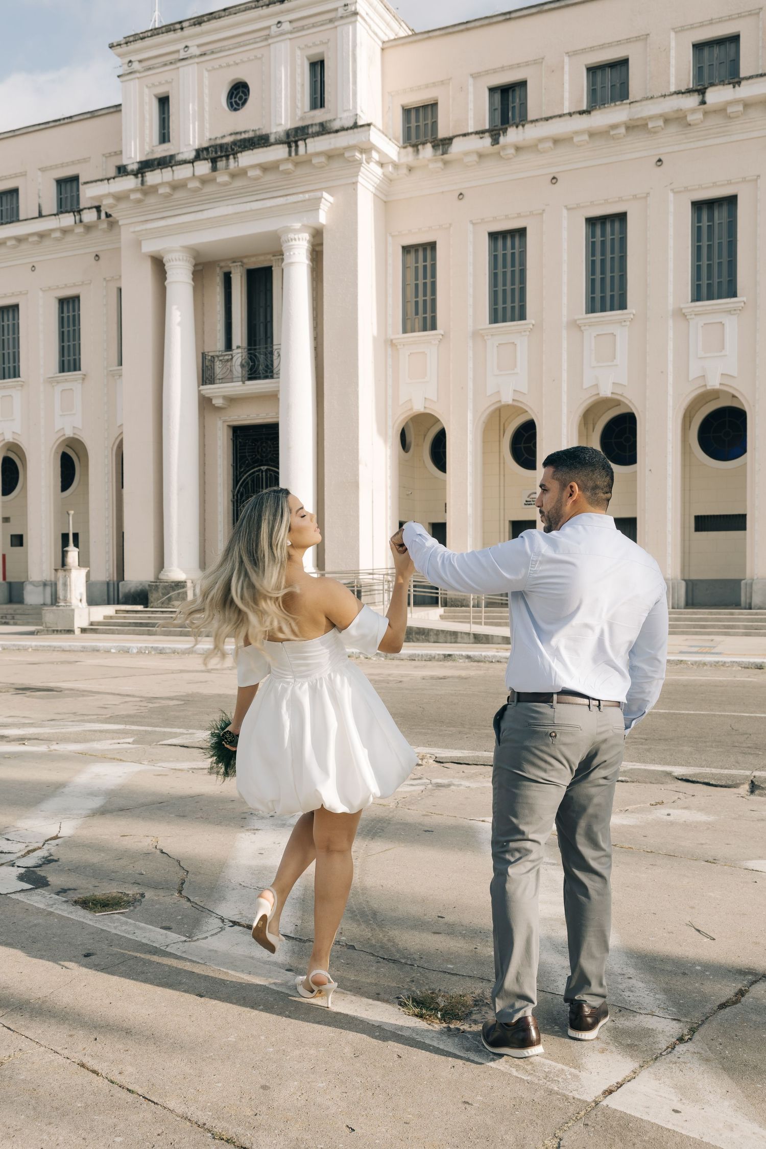 A couple in formal attire dances together outside a grand white neoclassical building with columns and arched windows.