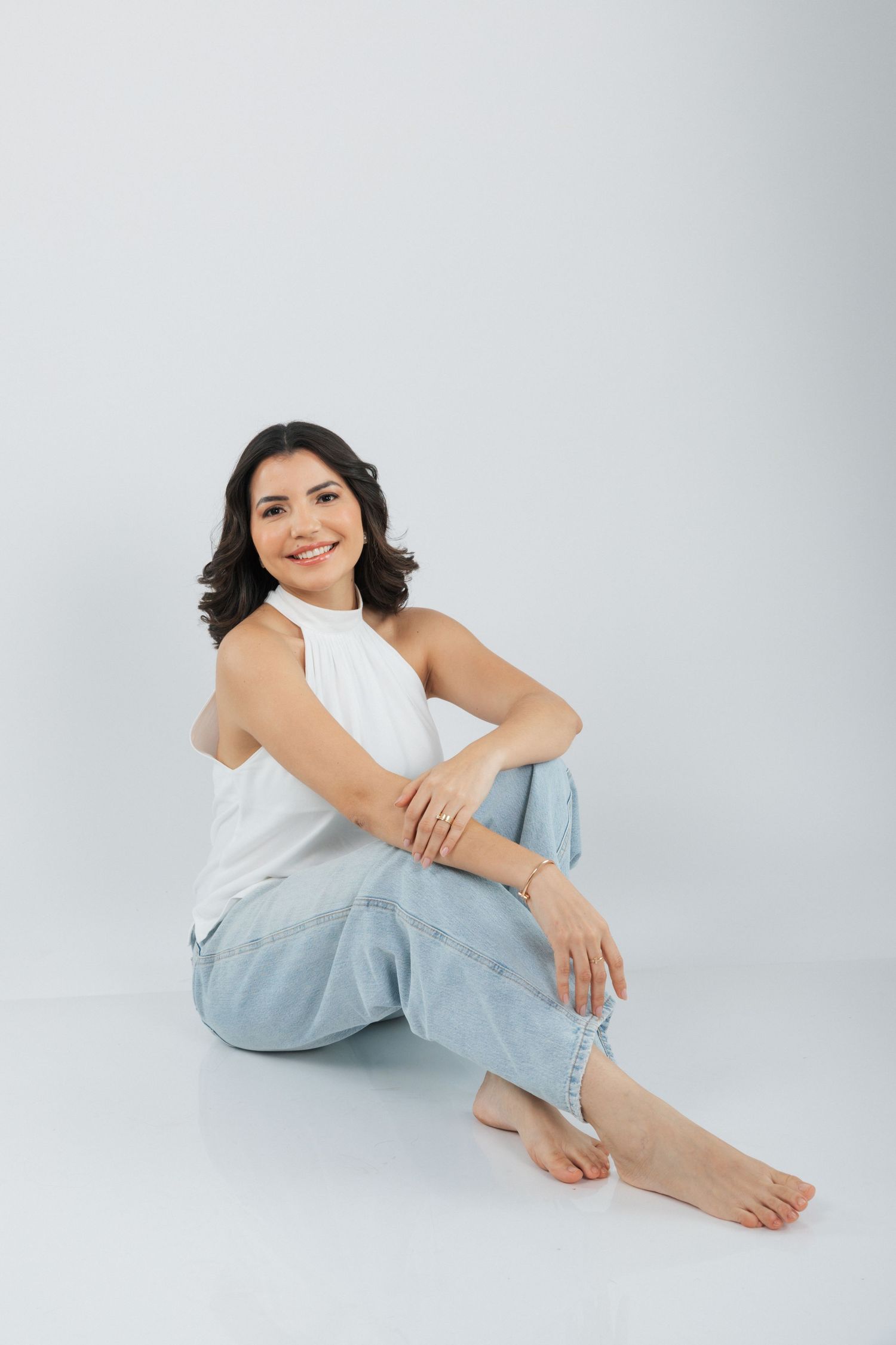 Casual portrait of a woman in a white sleeveless top and light jeans sitting barefoot against a gray background.
