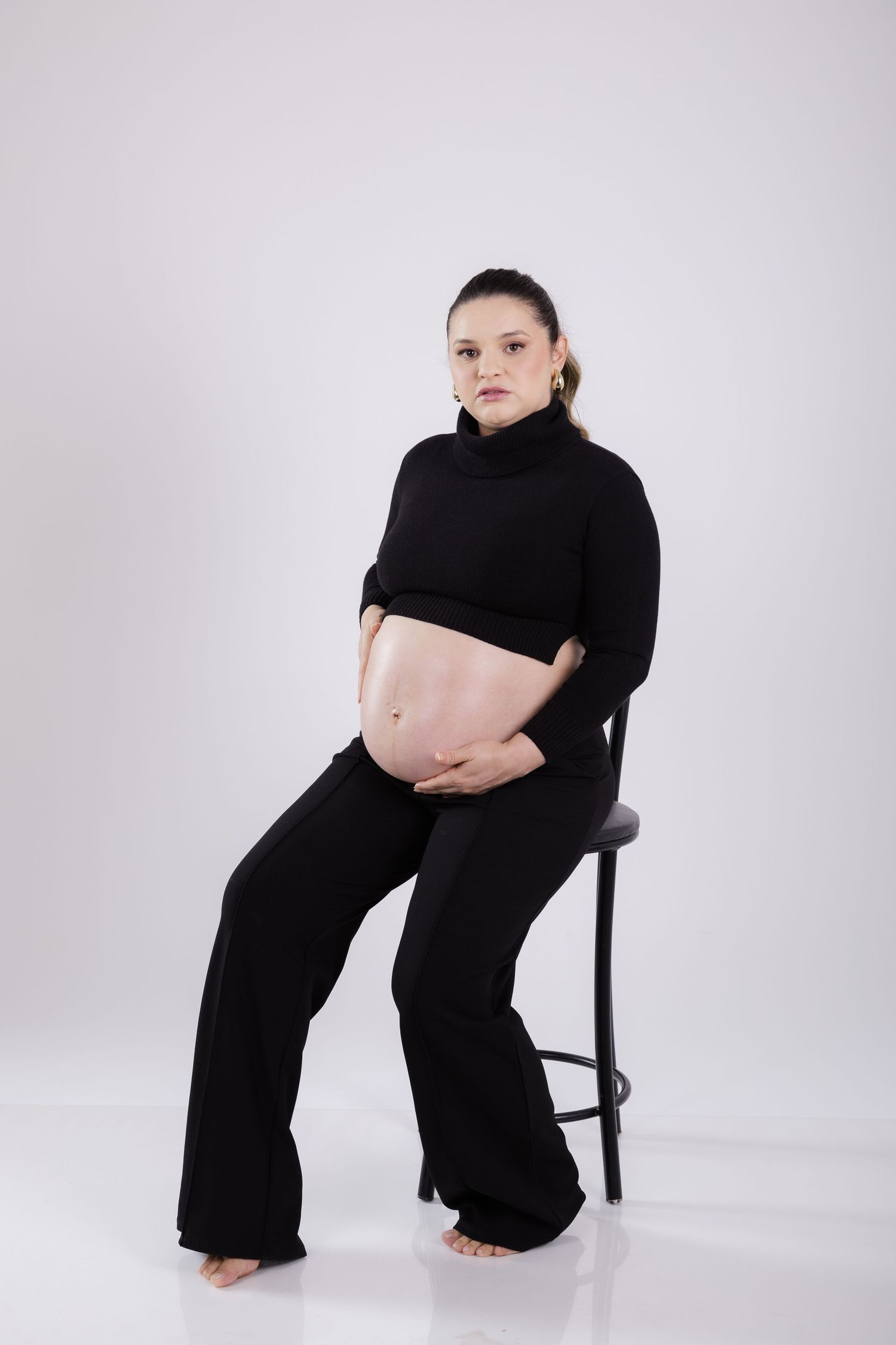 A pregnant person in black turtleneck crop top and pants sits on a stool against light gray studio backdrop.