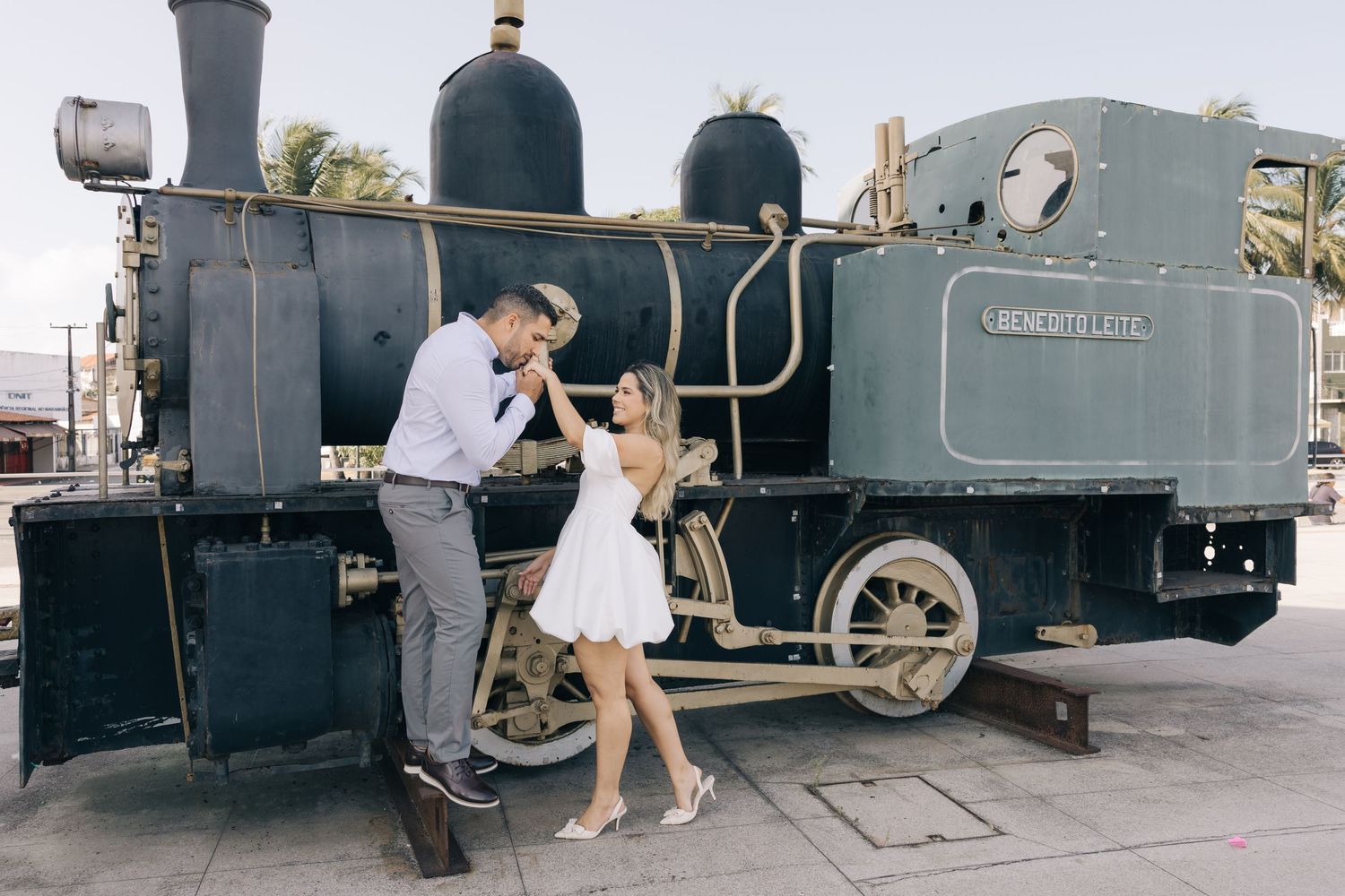 A vintage steam locomotive provides a nostalgic backdrop as two people interact near its wheels.