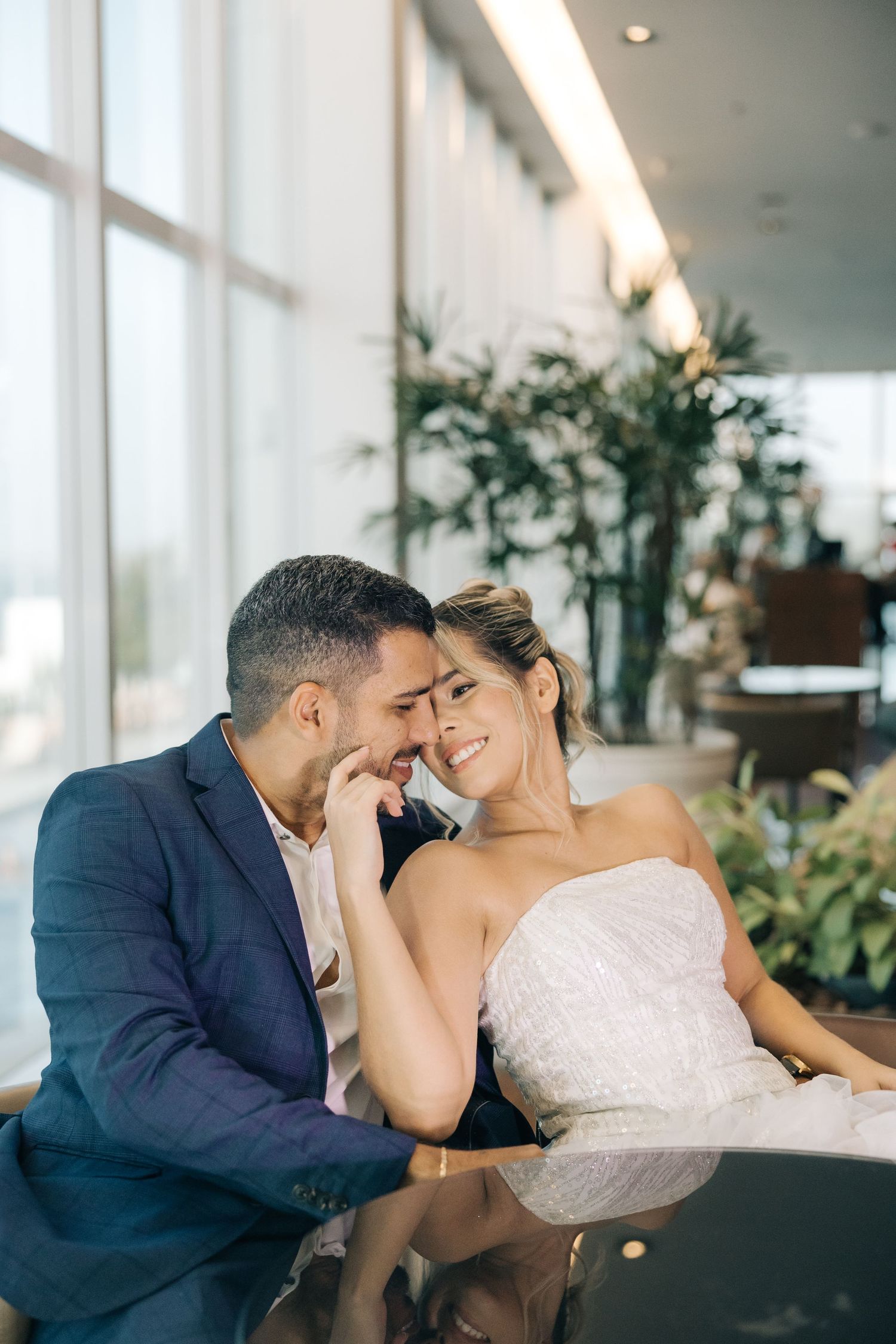 A bride in white gown and groom in navy suit share an intimate moment beside bright windows and indoor plants.