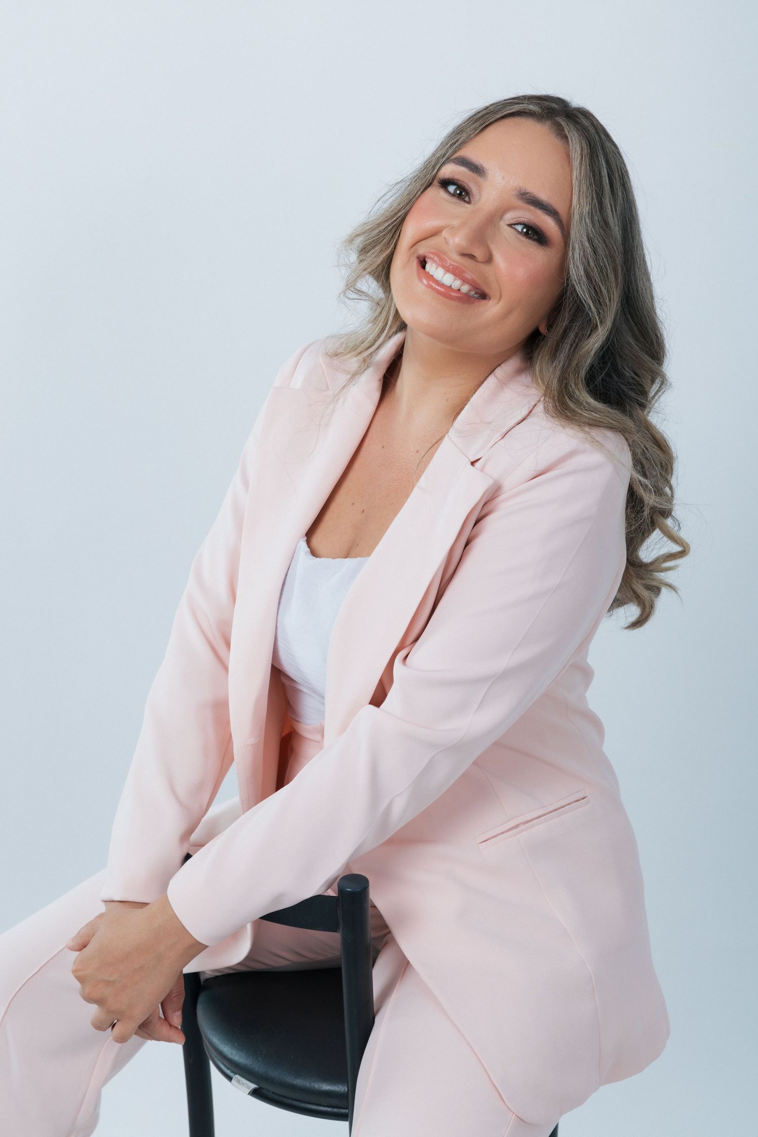 Professional portrait of someone wearing a soft pink blazer and white top while leaning on a chair against white background.