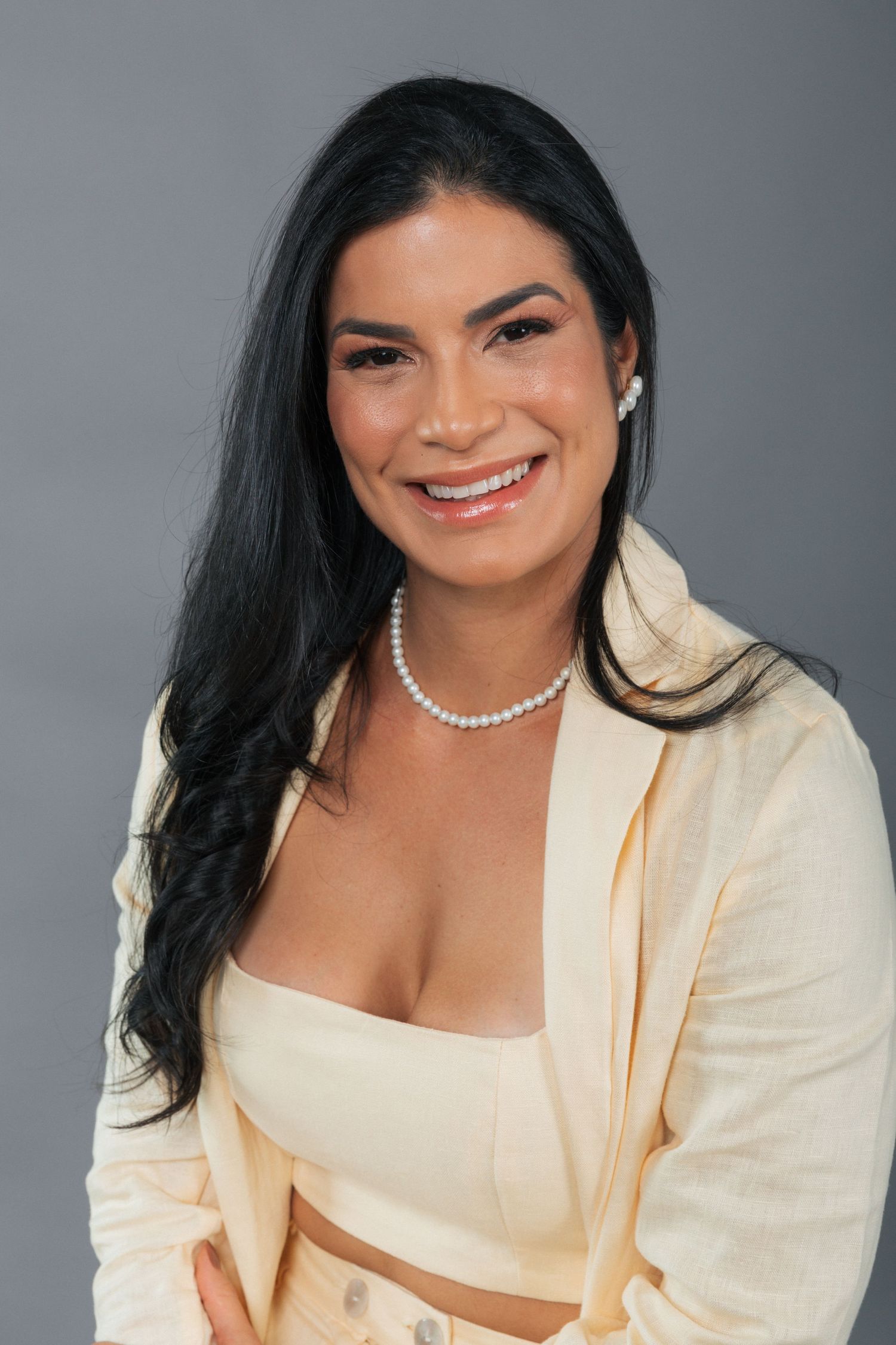 Professional studio portrait showing a person in a cream blazer and crop top with pearl necklace against grey background.