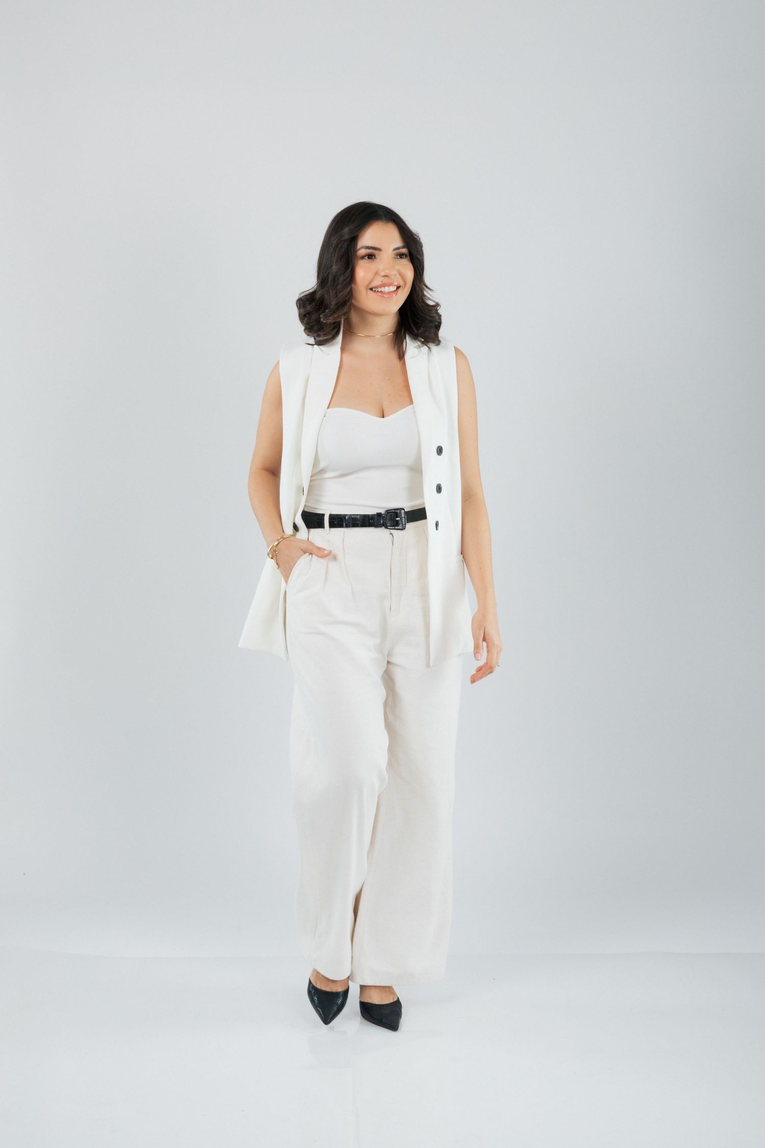 Full body portrait in crisp white pantsuit with black belt and heels against light background.