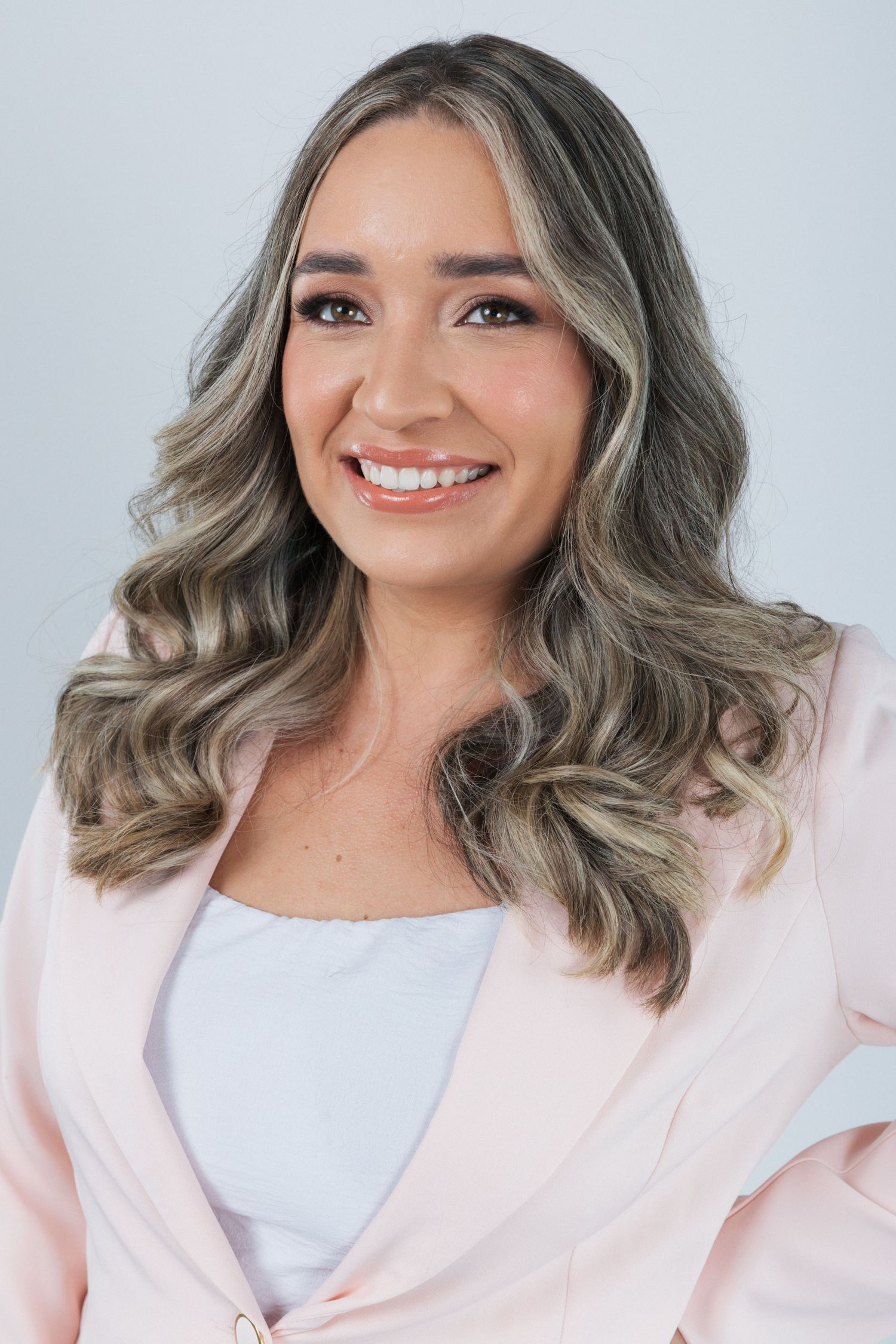 Professional portrait of someone with wavy silver-blonde hair wearing a pink blazer and white top against a light background.