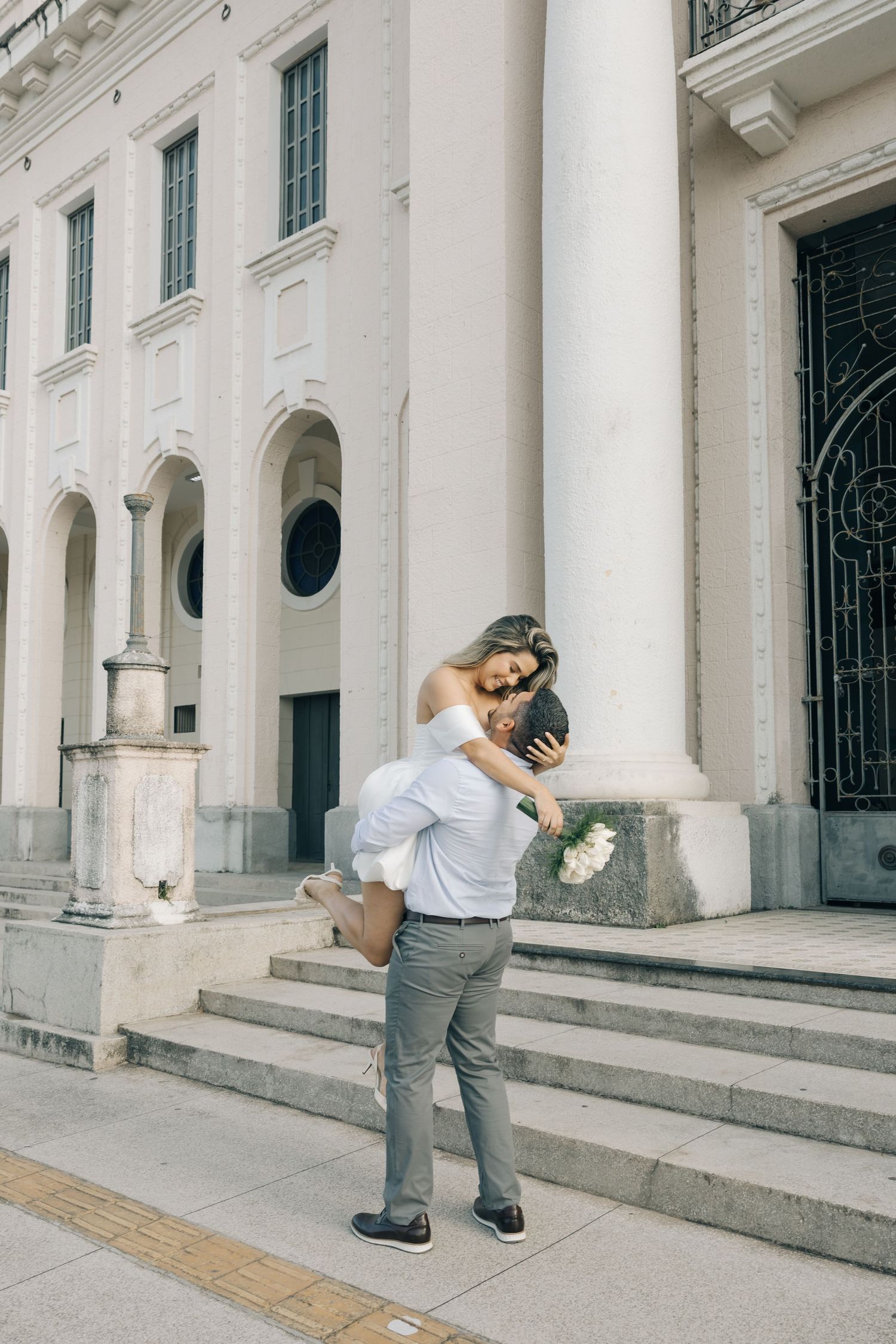 A joyful romantic moment on the steps of a grand white building with classical columns and architectural details.
