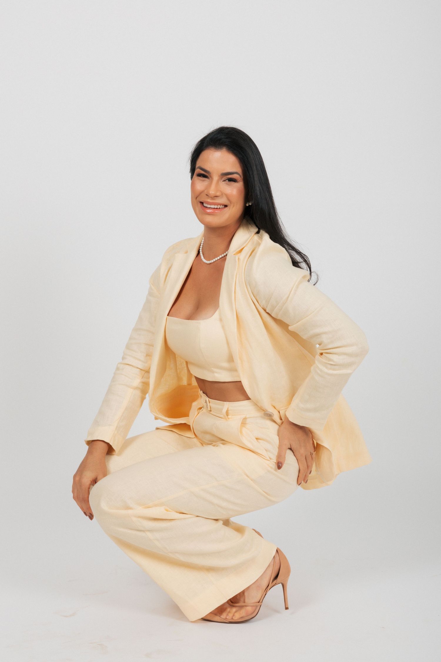 Professional studio portrait of someone in a cream colored pantsuit with matching crop top kneeling against white backdrop.