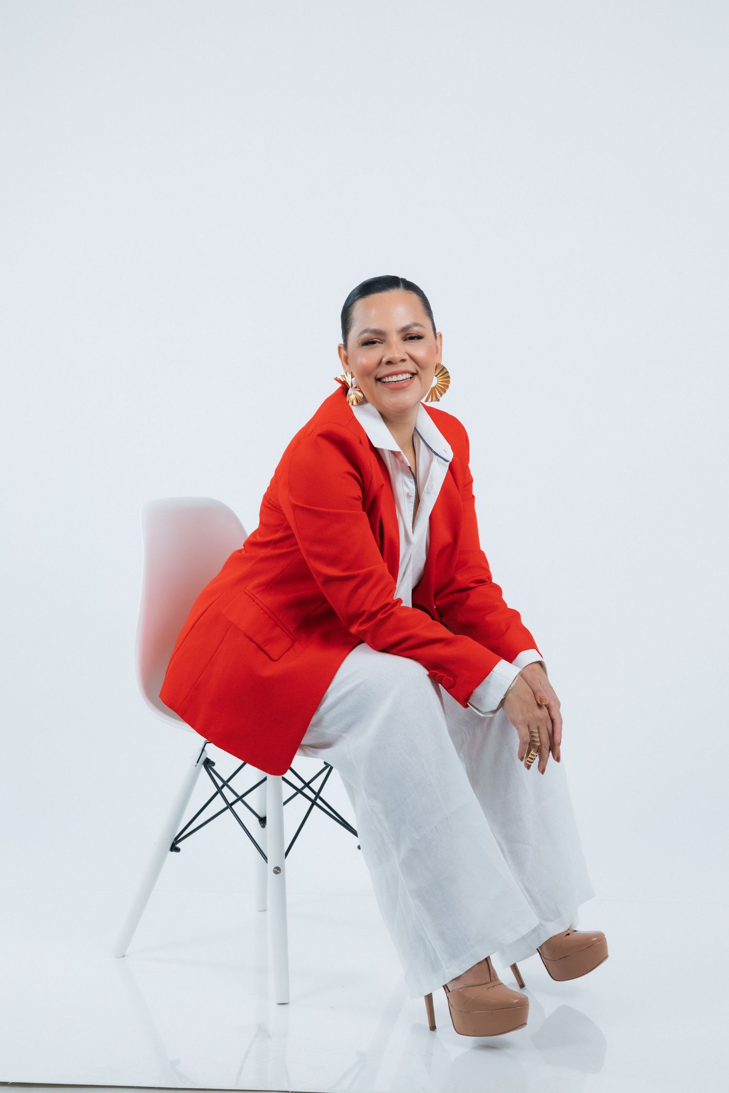 Stylish woman in red blazer and white pants seated on modern chair against plain background.