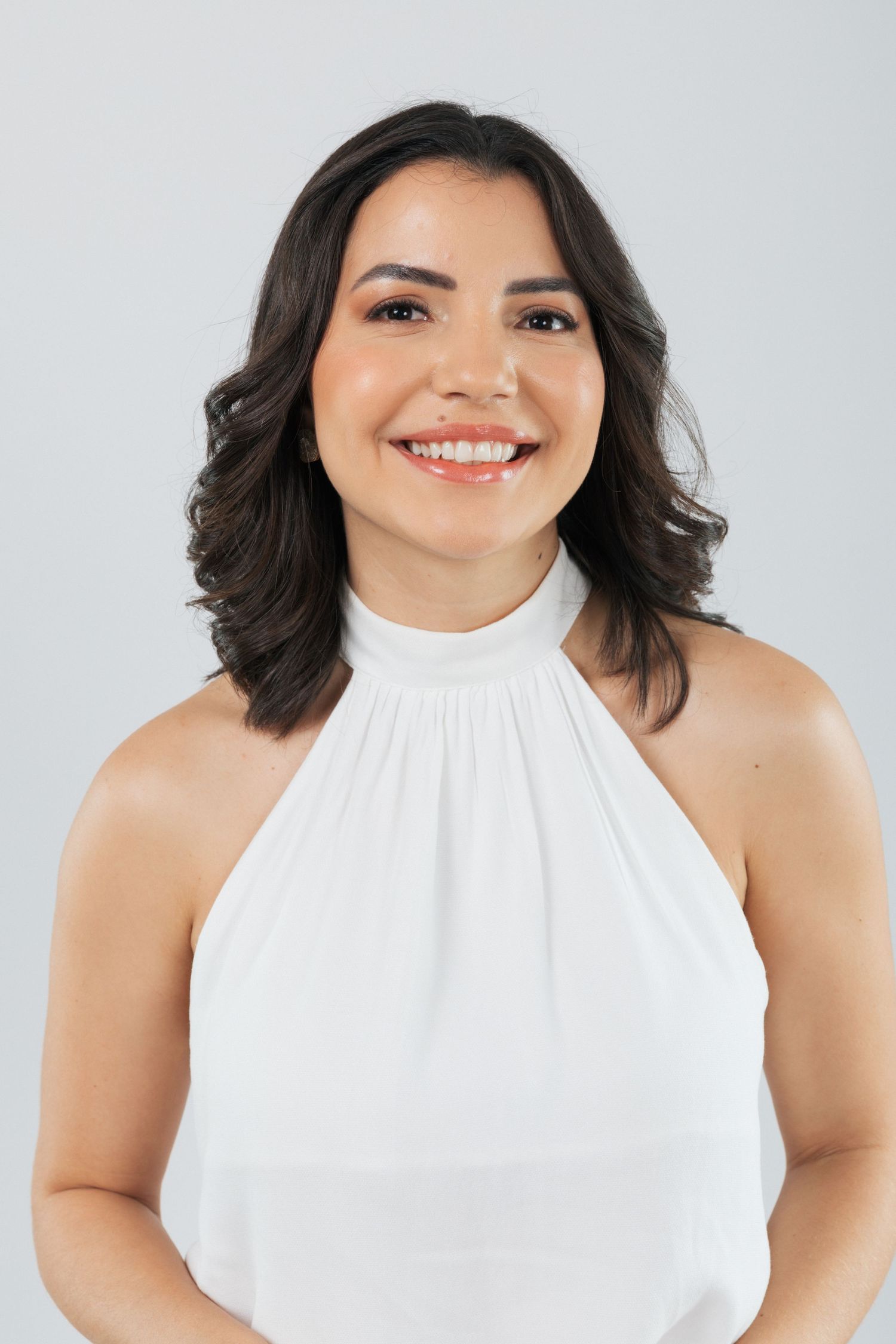 Professional headshot of a woman in a white halter top smiling warmly against a light gray background.