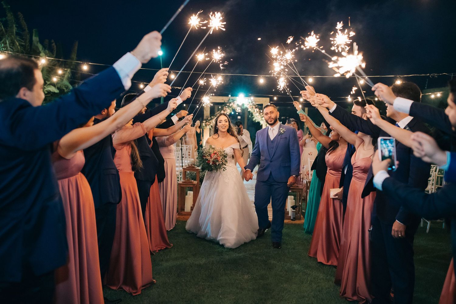 Wedding party holds sparklers creating a magical archway during an outdoor evening celebration on a grass lawn.