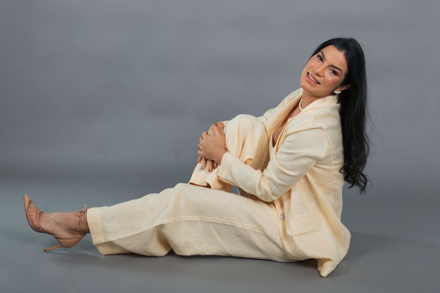 Person in cream colored linen suit lounges expressively on gray background, creating artistic fashion pose.
