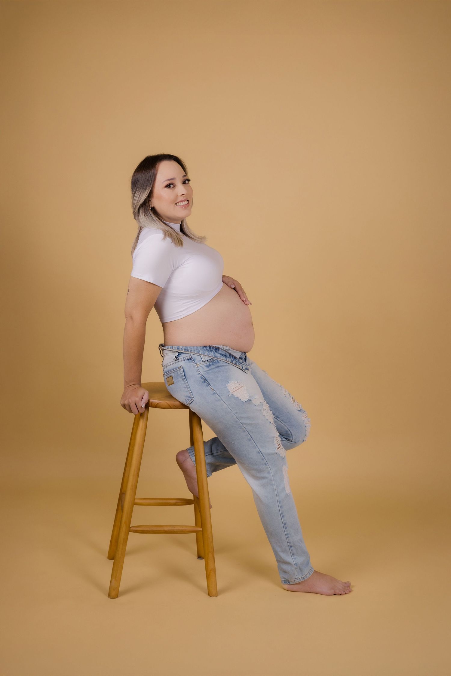 Woman in white crop top and distressed jeans sits sideways on wooden stool in studio setting.