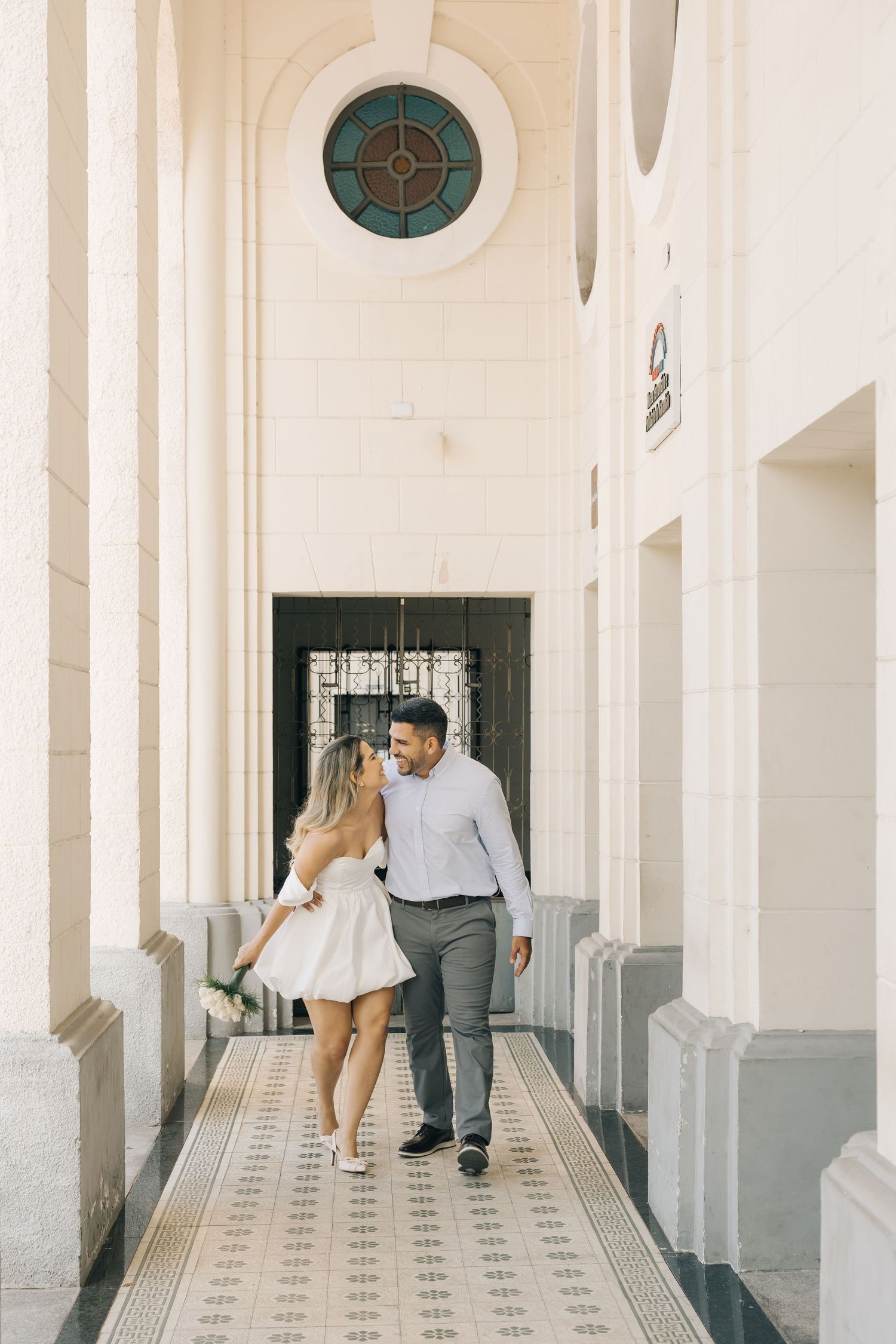 A couple walks together in a vintage hallway with ornate tiled floors and circular window features.