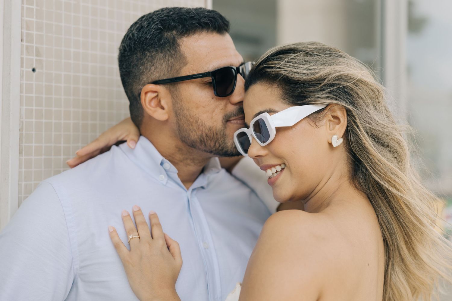 A stylish couple wearing sunglasses embrace in a sun-lit hallway while sharing an intimate moment.