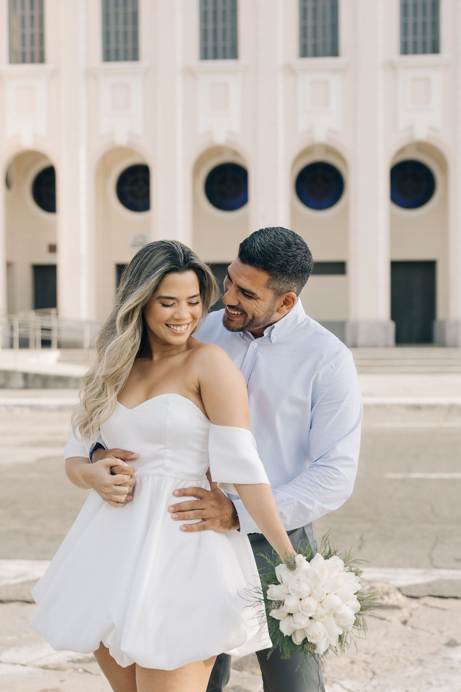 A romantic couple embraces near a classical building with white columns and circular windows, with white flowers in hand.
