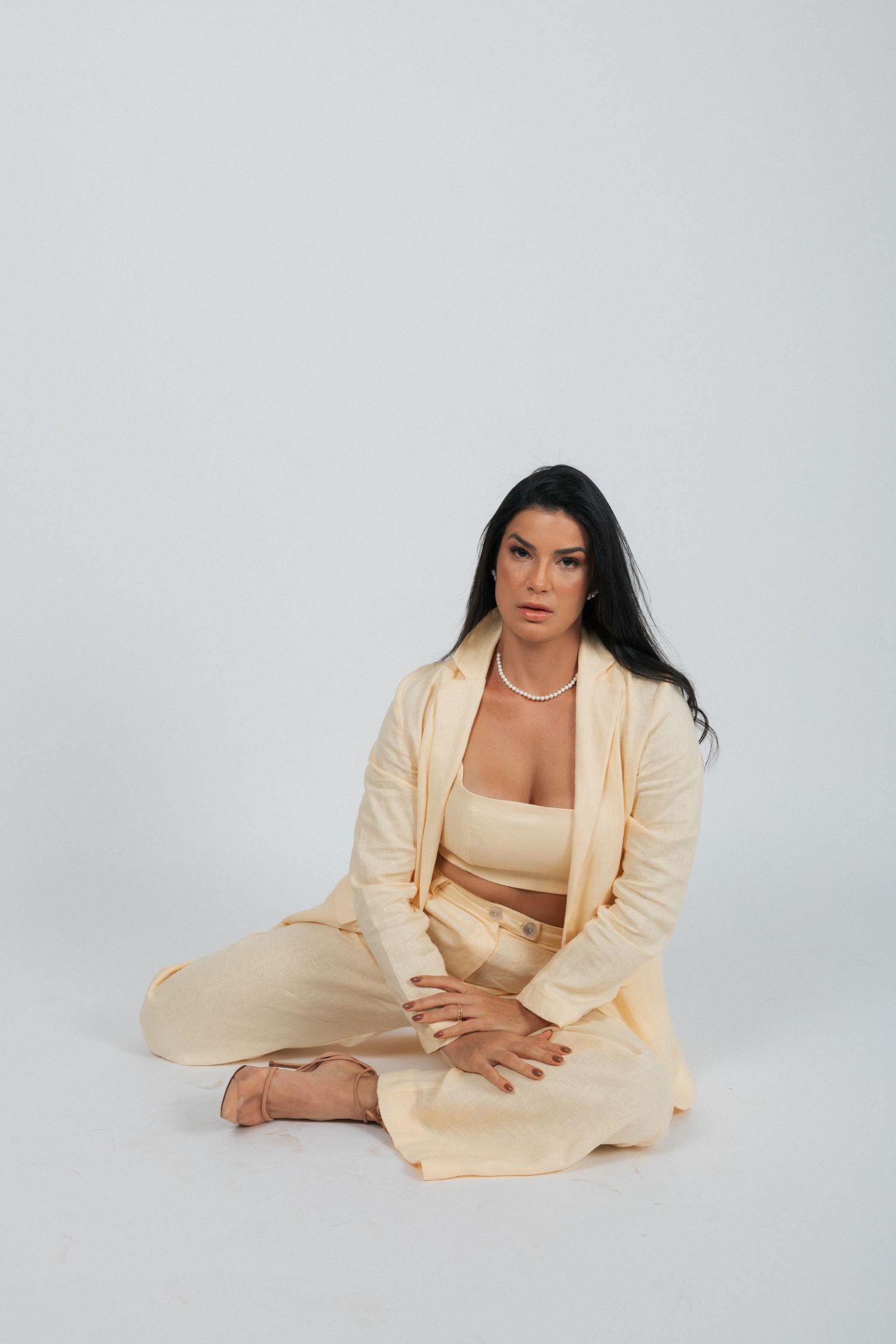 Woman in cream-colored loungewear set sits cross-legged on floor wearing pearl necklace.