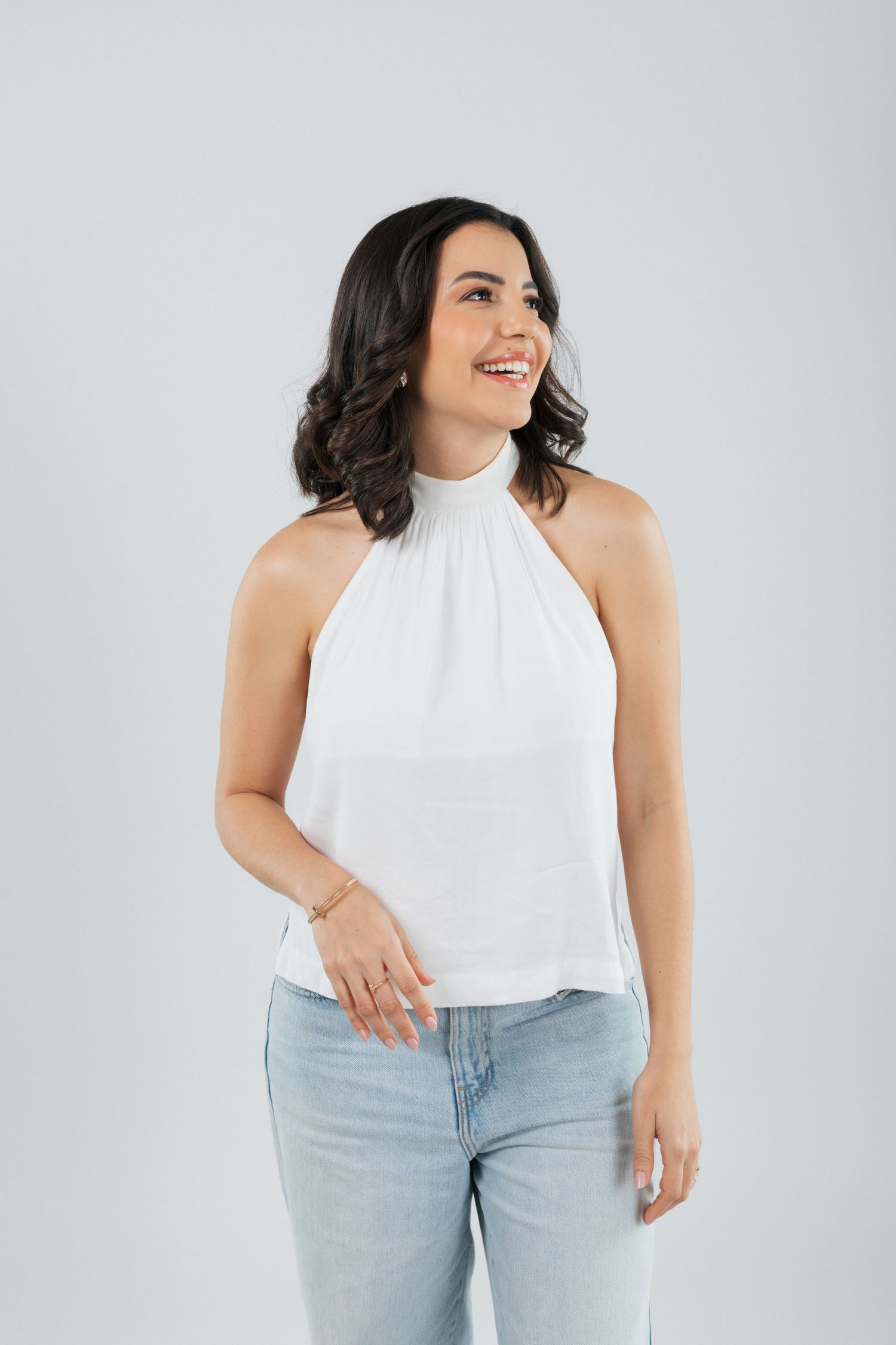 Woman in casual white sleeveless top and jeans smiles while posing against neutral background.