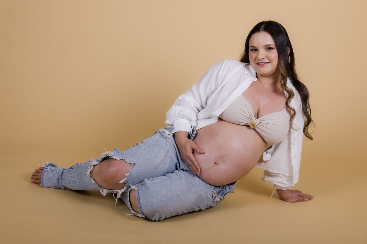 Expectant mother in white shirt and ripped jeans reclines on tan backdrop during maternity photoshoot.