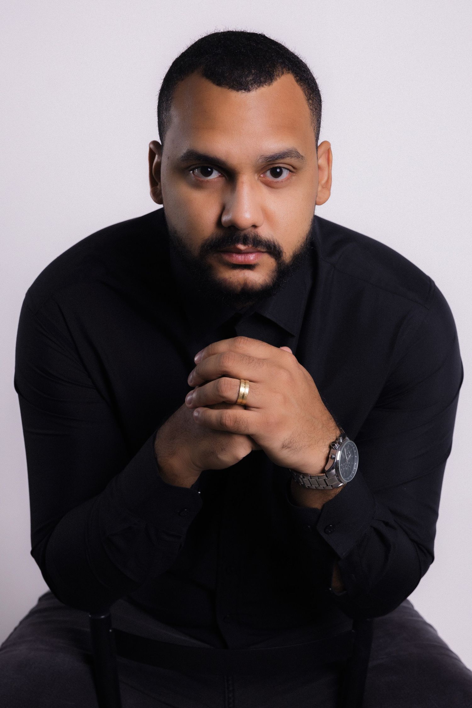 Professional studio portrait of someone in a black sweater seated against light background wearing a watch and ring.