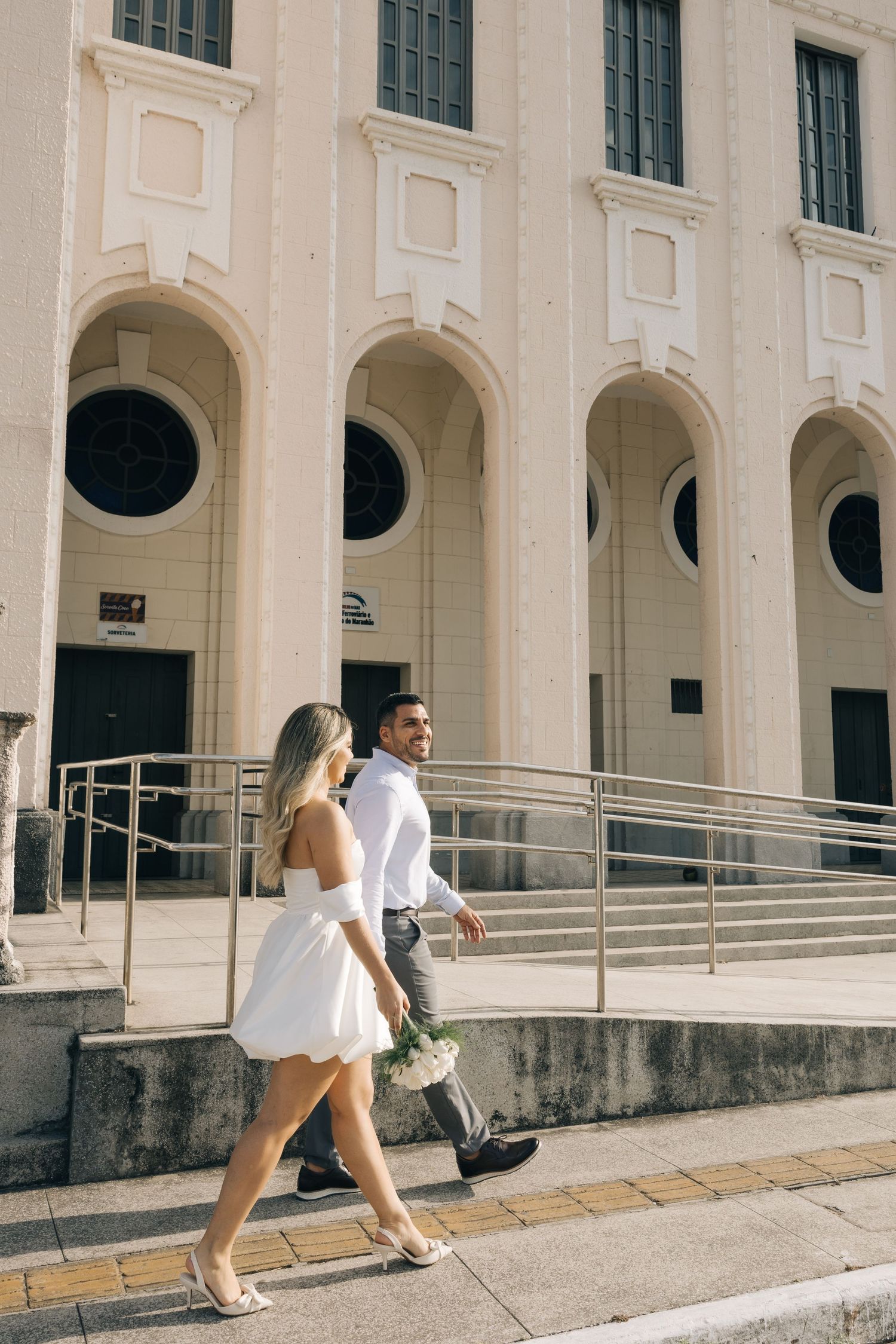 A couple in formal attire walking along a sidewalk next to a classic building facade.