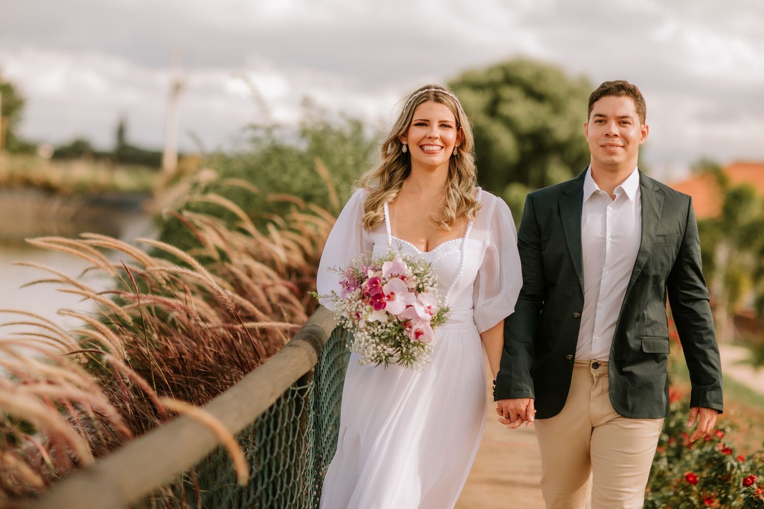 A joyful couple walks together on a boardwalk surrounded by pampas grass and flowers during a seaside wedding.