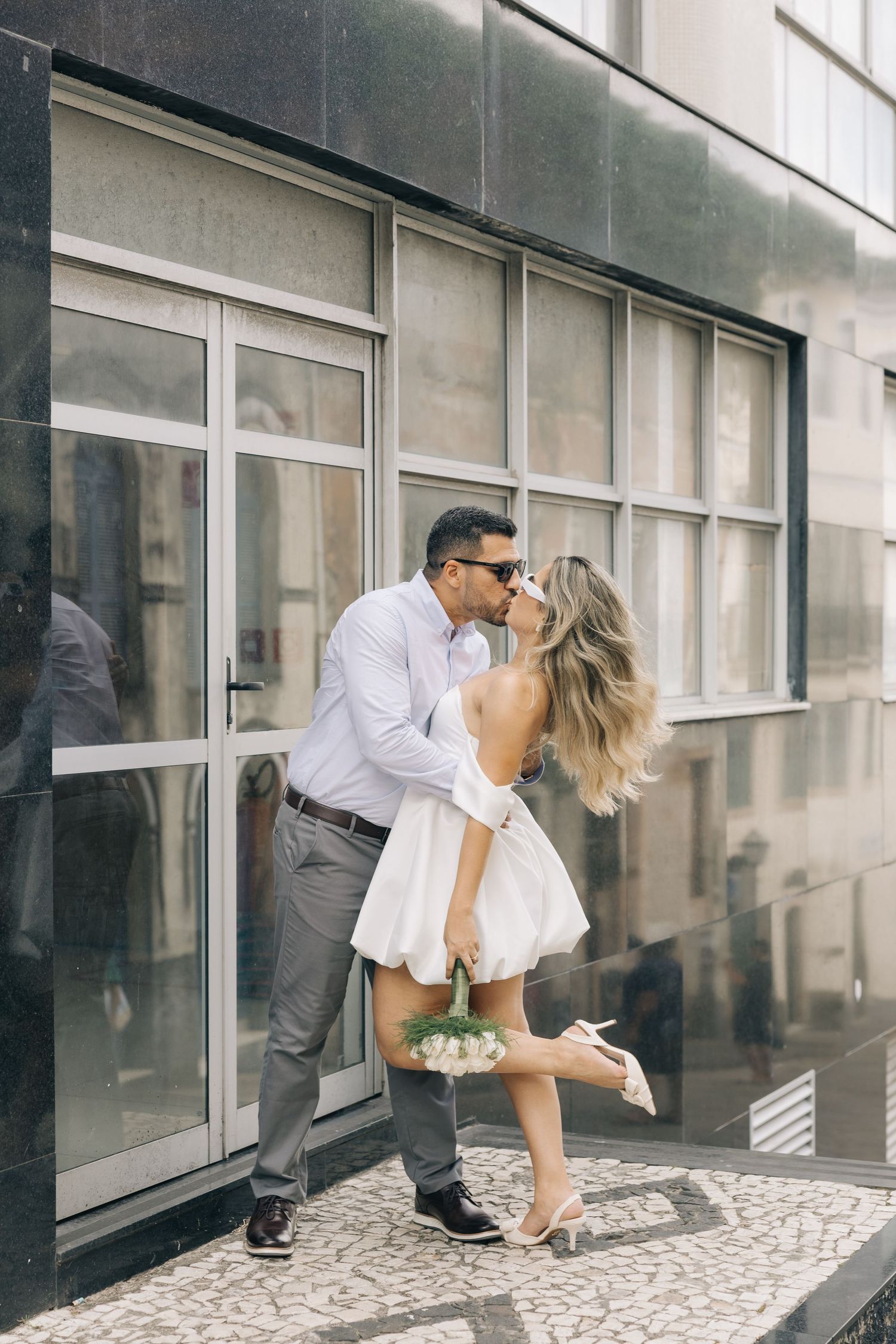 A romantic moment captured as a couple shares a kiss outside a modern building with large windows.