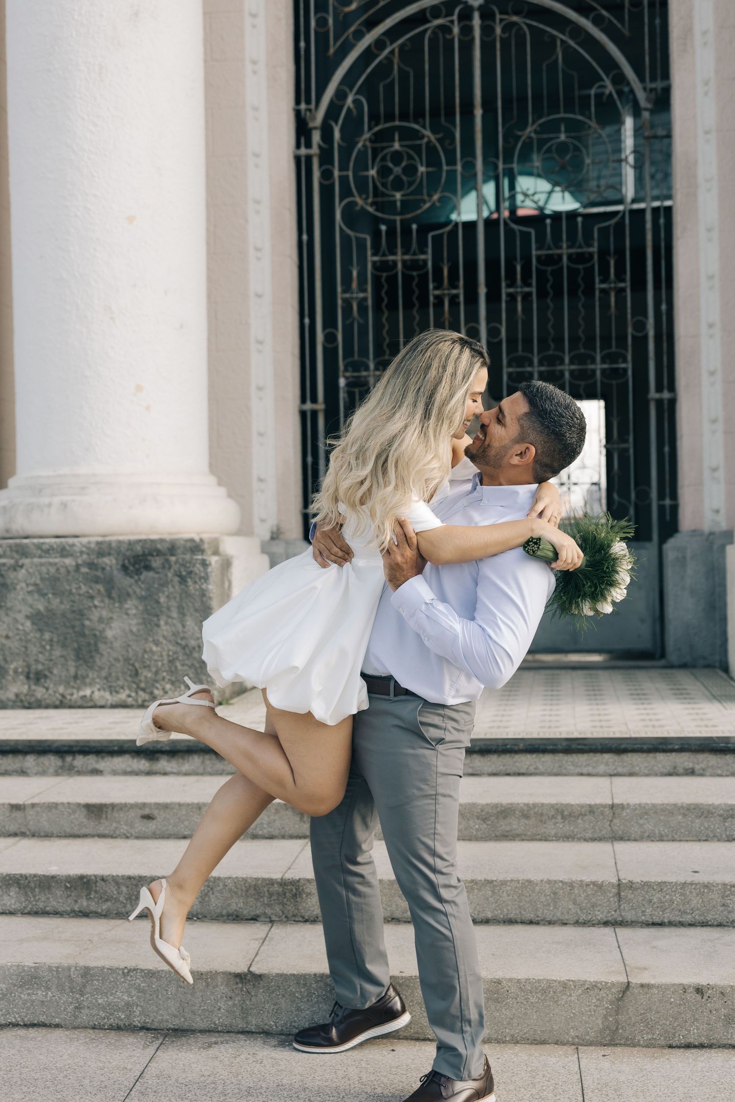 A romantic couple embraces on church steps in front of an ornate iron gate, with the woman in a white dress leaping up.