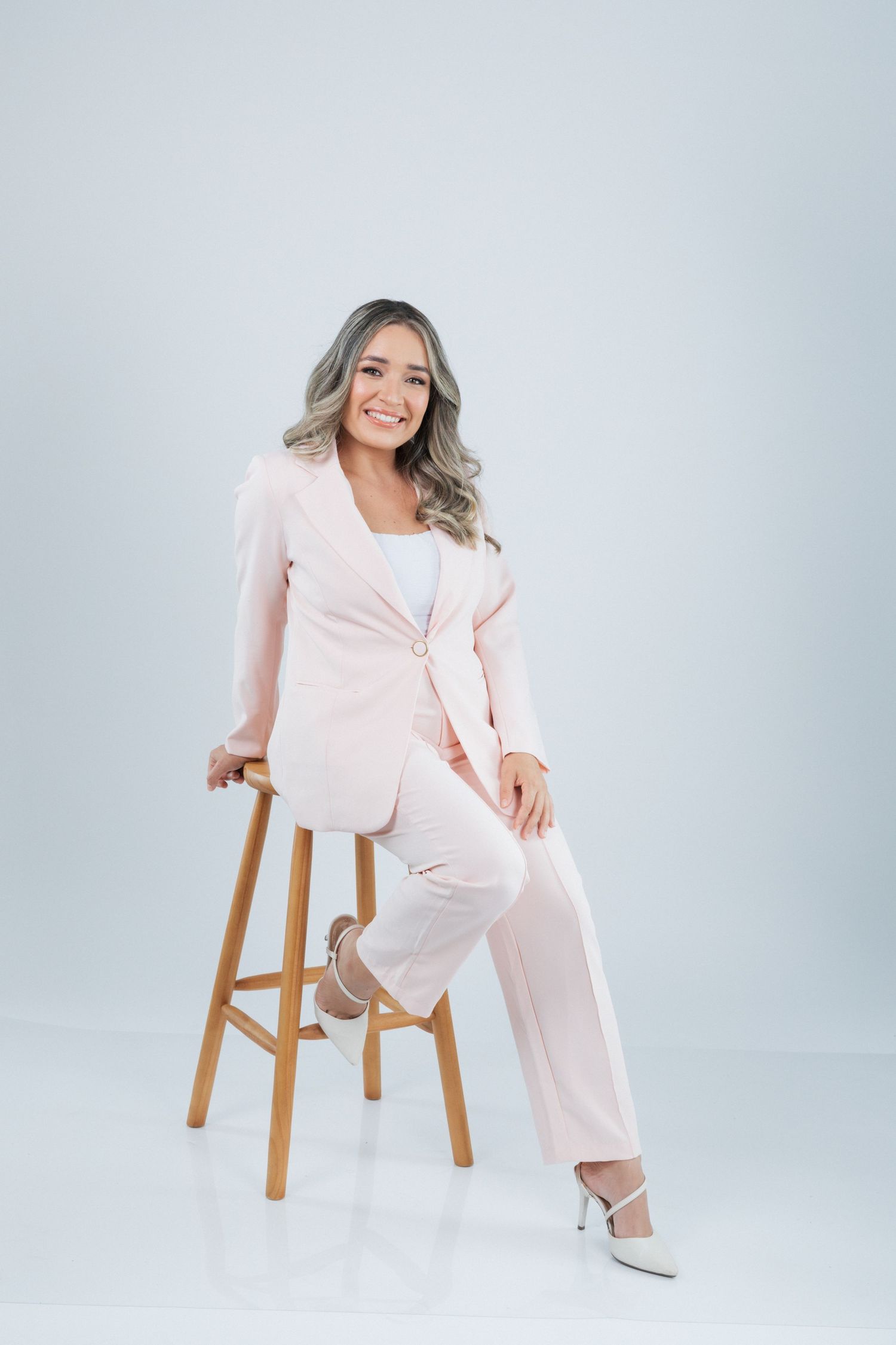 Person in soft pink business suit sits elegantly on wooden stool against light gray backdrop, displaying professional style.