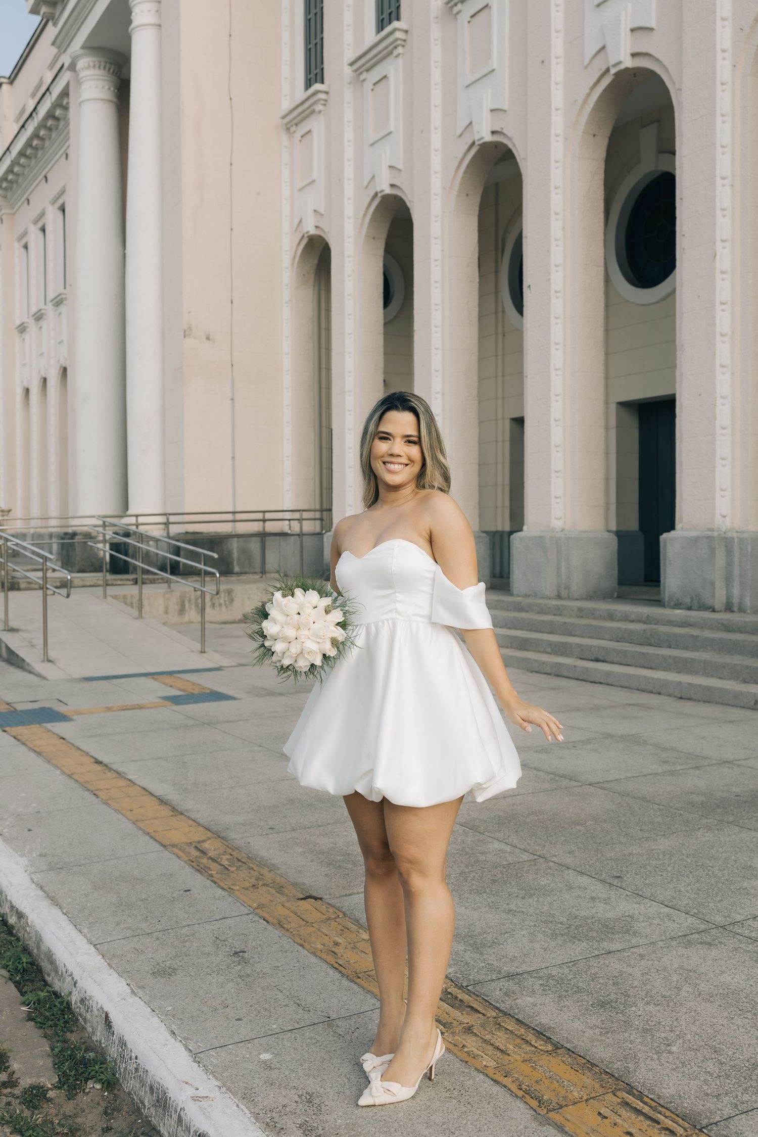 A bride in a short white off-shoulder dress and heels poses with a white bouquet outside a classical building with arches.