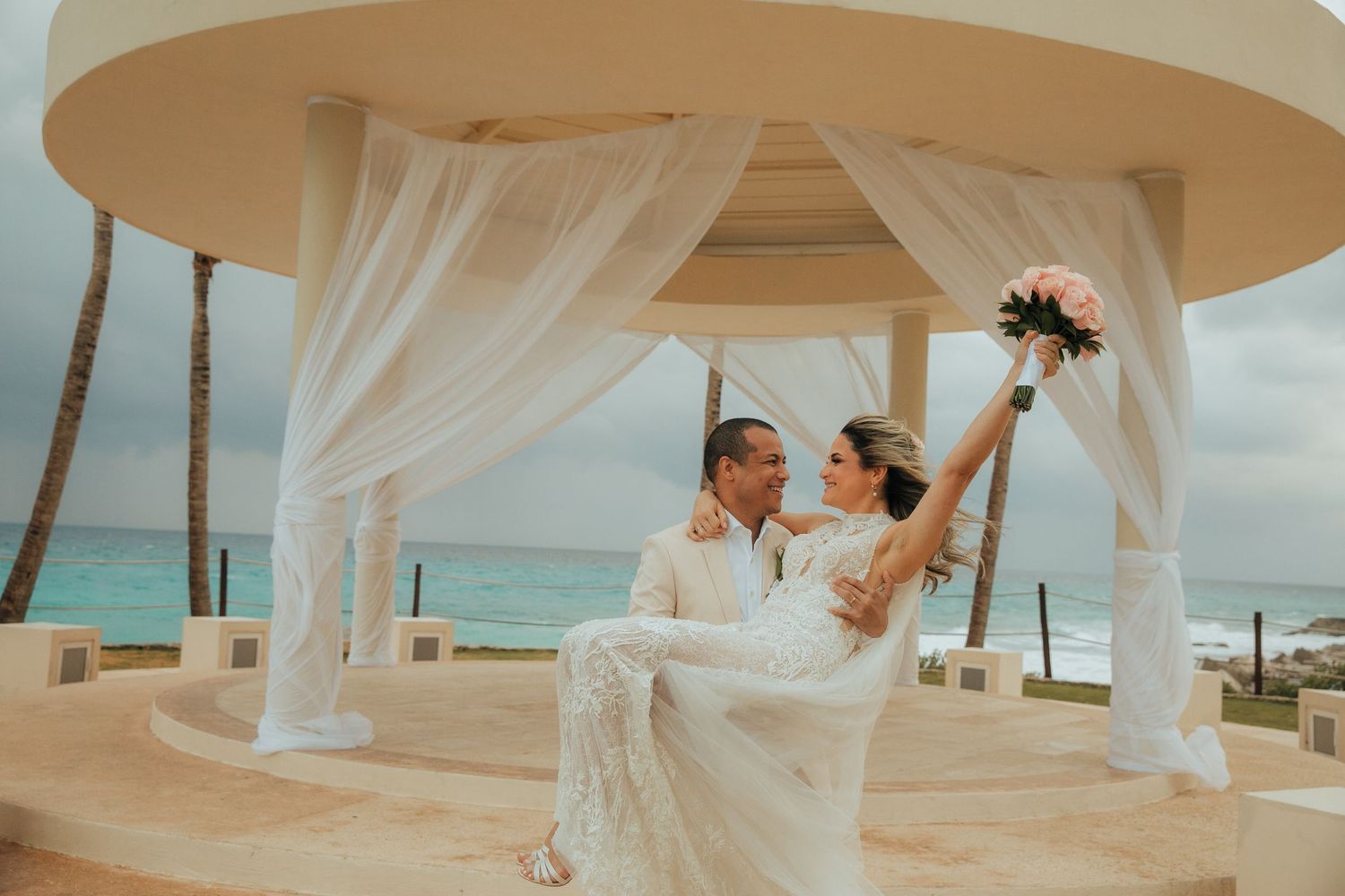 A couple celebrates their wedding day beneath a draped gazebo overlooking turquoise ocean waters on a beach.
