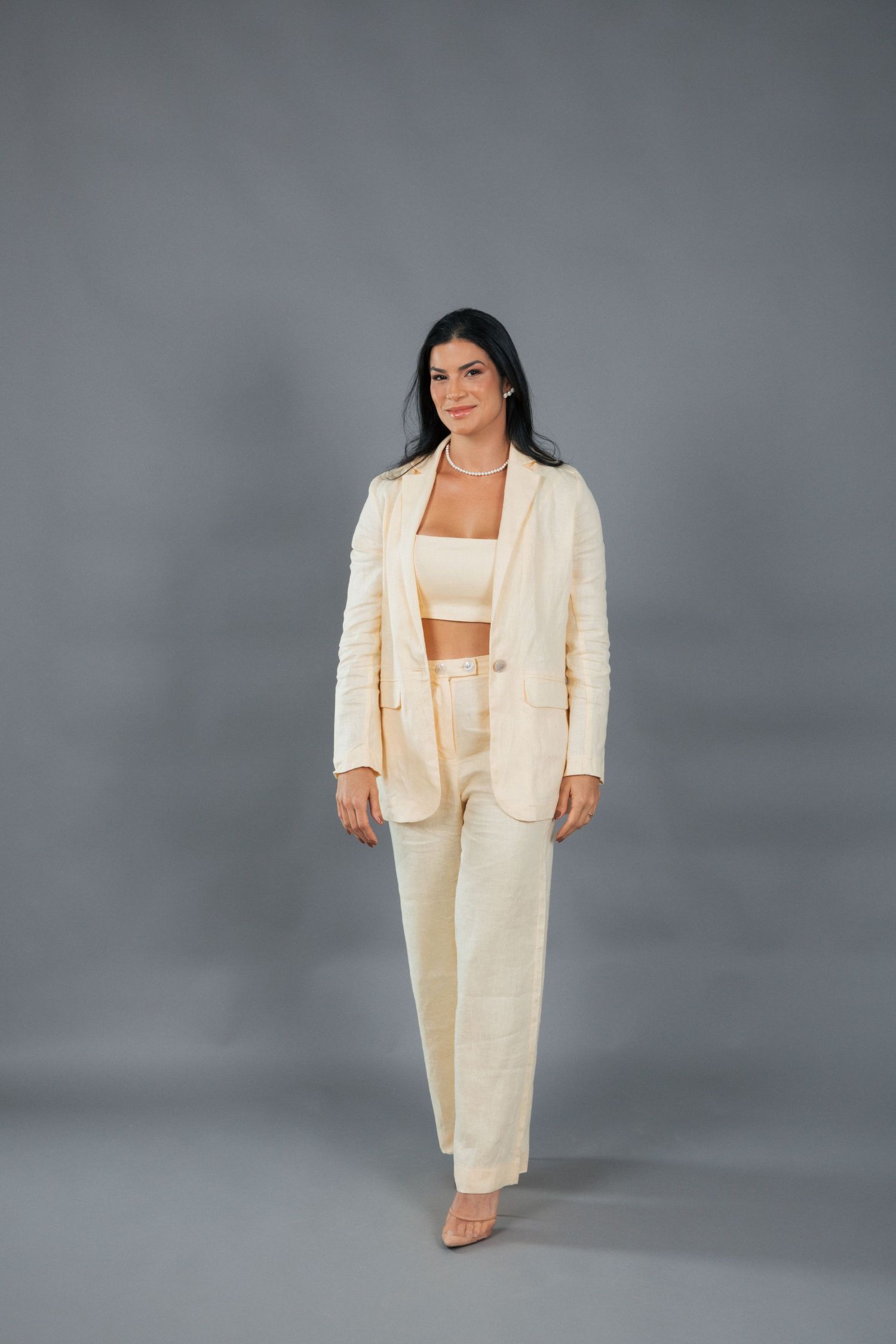 A person models a cream-colored power suit with a matching crop top and pants against a gray studio background.