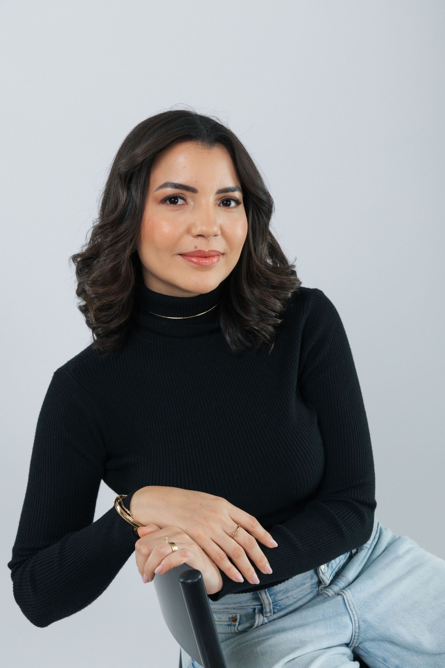 Modern studio portrait in black turtleneck and light jeans on chair against gray backdrop.