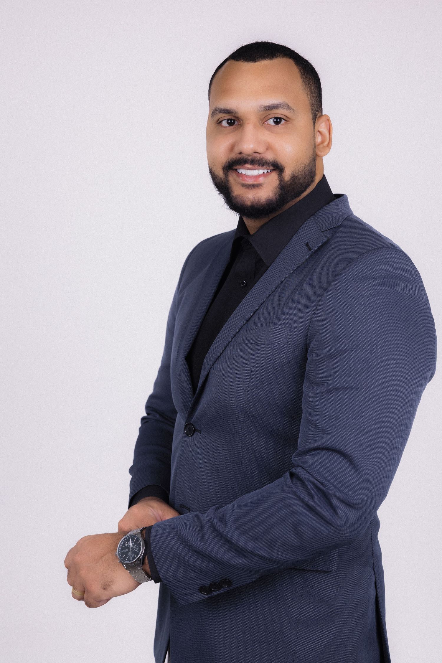 Professional business portrait of a smiling person wearing a navy suit and black shirt against a light background.