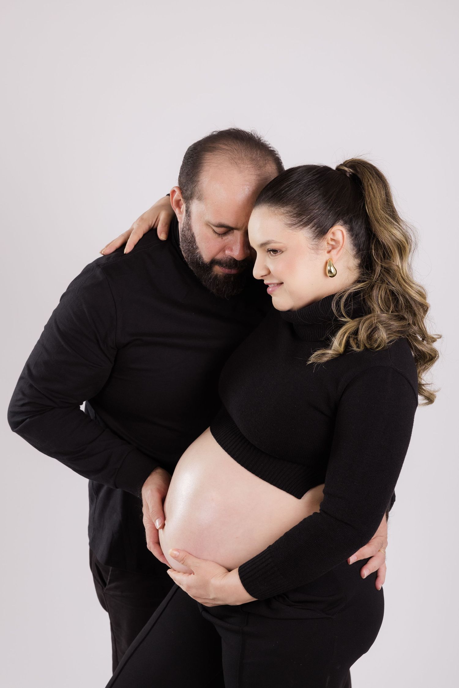 Maternity photo of a couple in black outfits embracing and touching a pregnant belly against a light background.