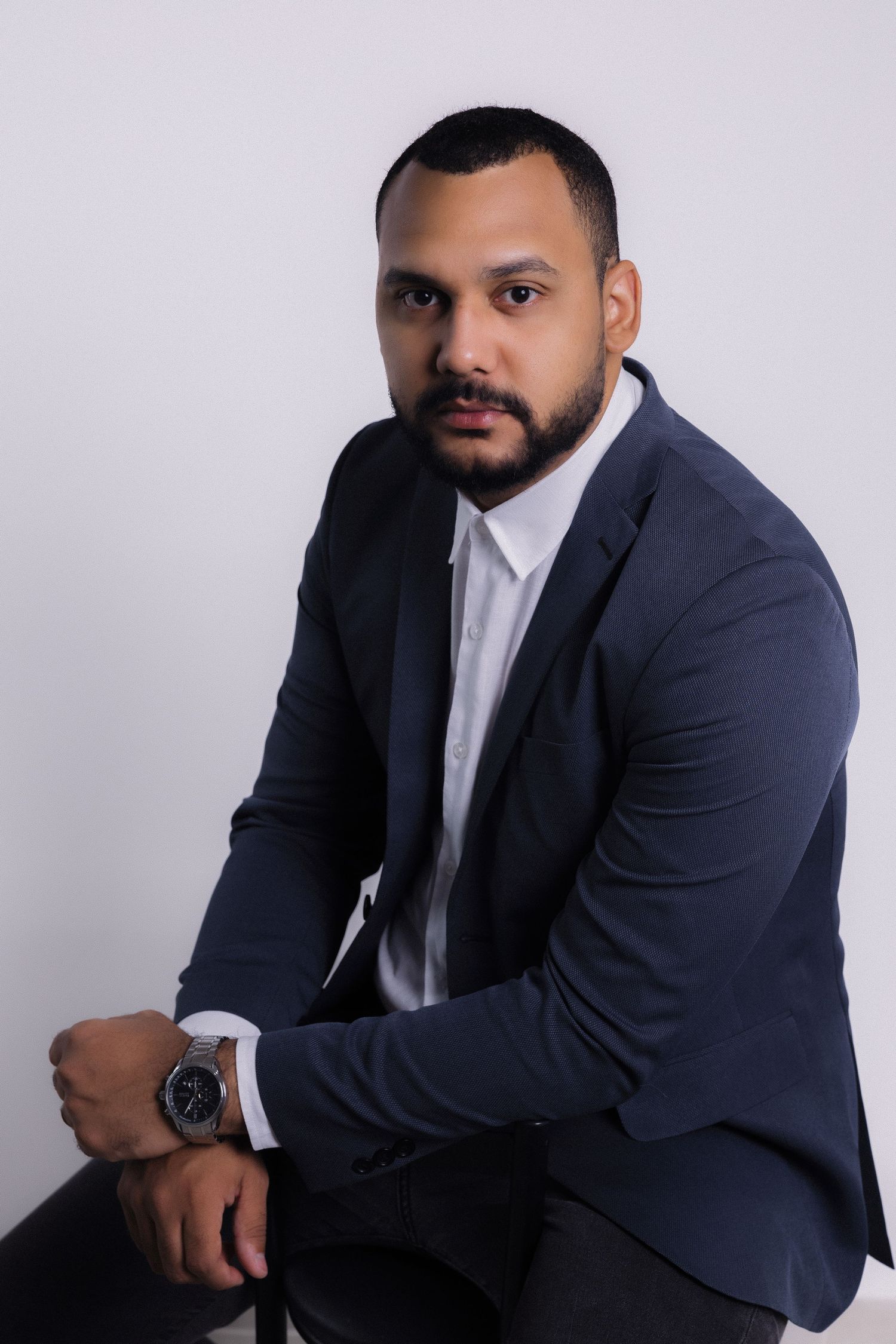 Professional portrait of someone wearing a navy blue suit and white dress shirt seated against a light gray background.