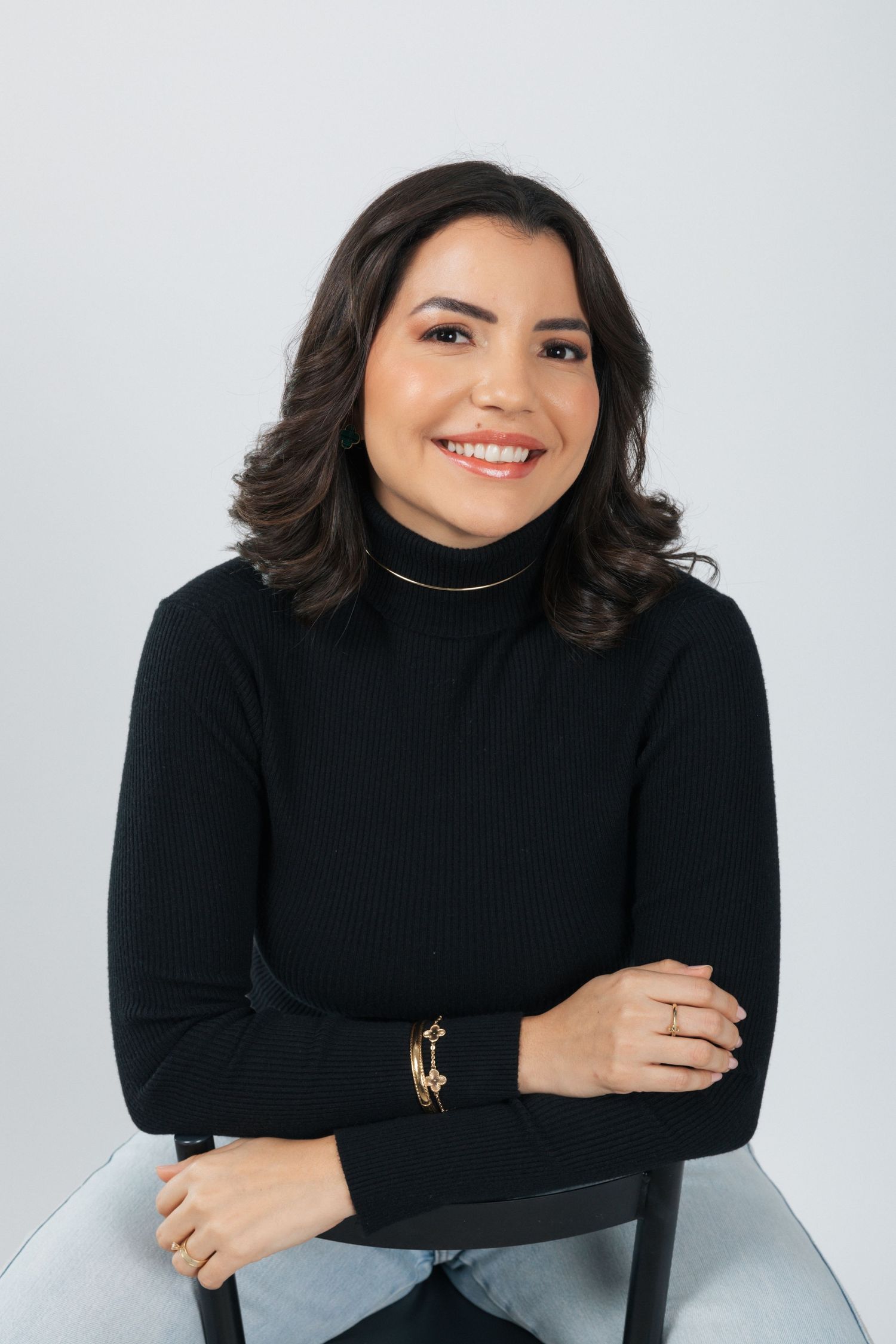 Professional headshot wearing a black turtleneck with gold bracelet against gray background.