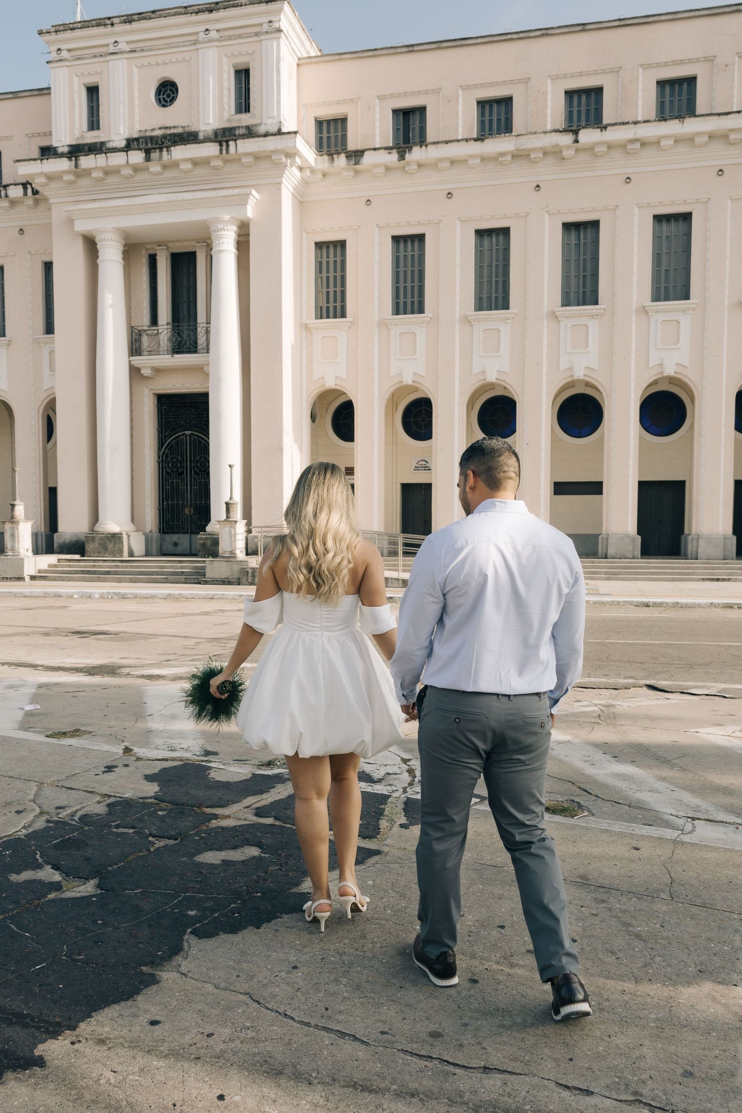 Couple walking towards a neoclassical building with white facade and circular windows on a sunny day.