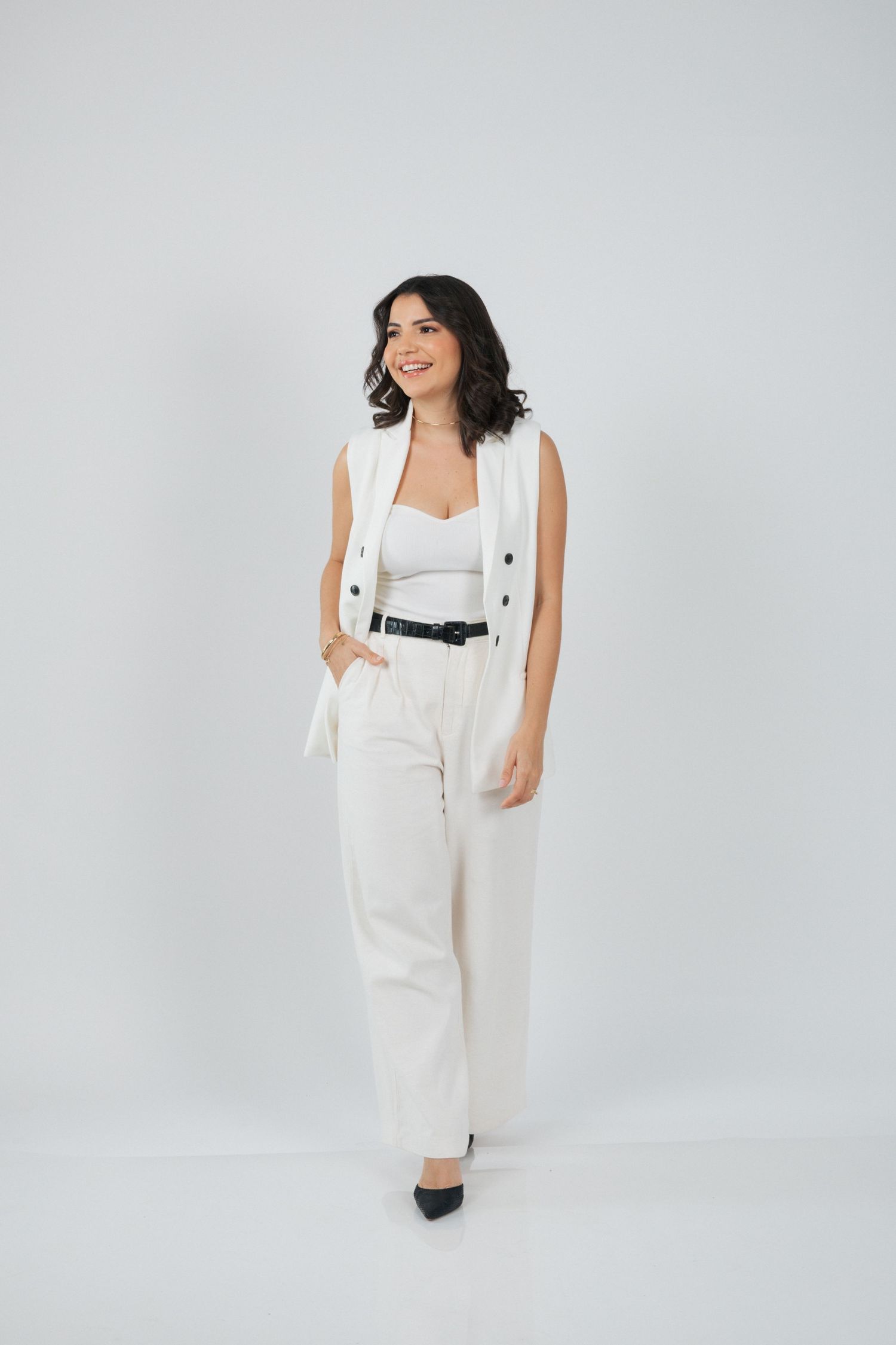 Stylish full-body shot in white pantsuit with black accessories against light backdrop.