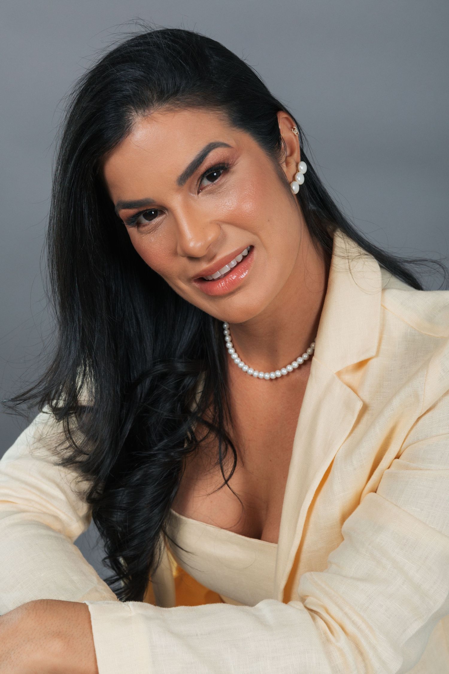 Professional portrait of someone in a cream blazer and pearl necklace with long dark hair against a gray background.