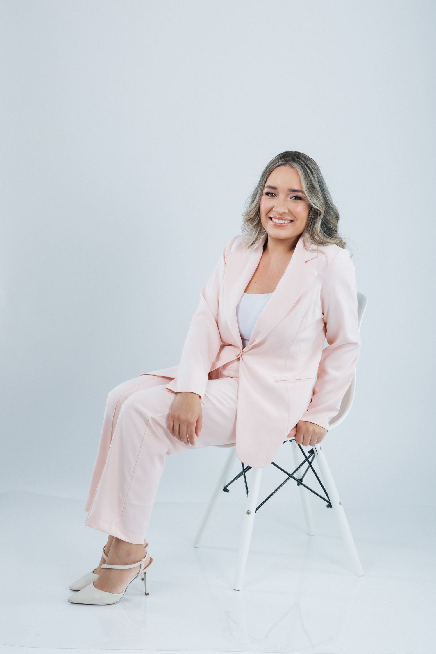 Someone in a pale pink pantsuit smiles warmly while sitting on a modern white chair in a studio setting.