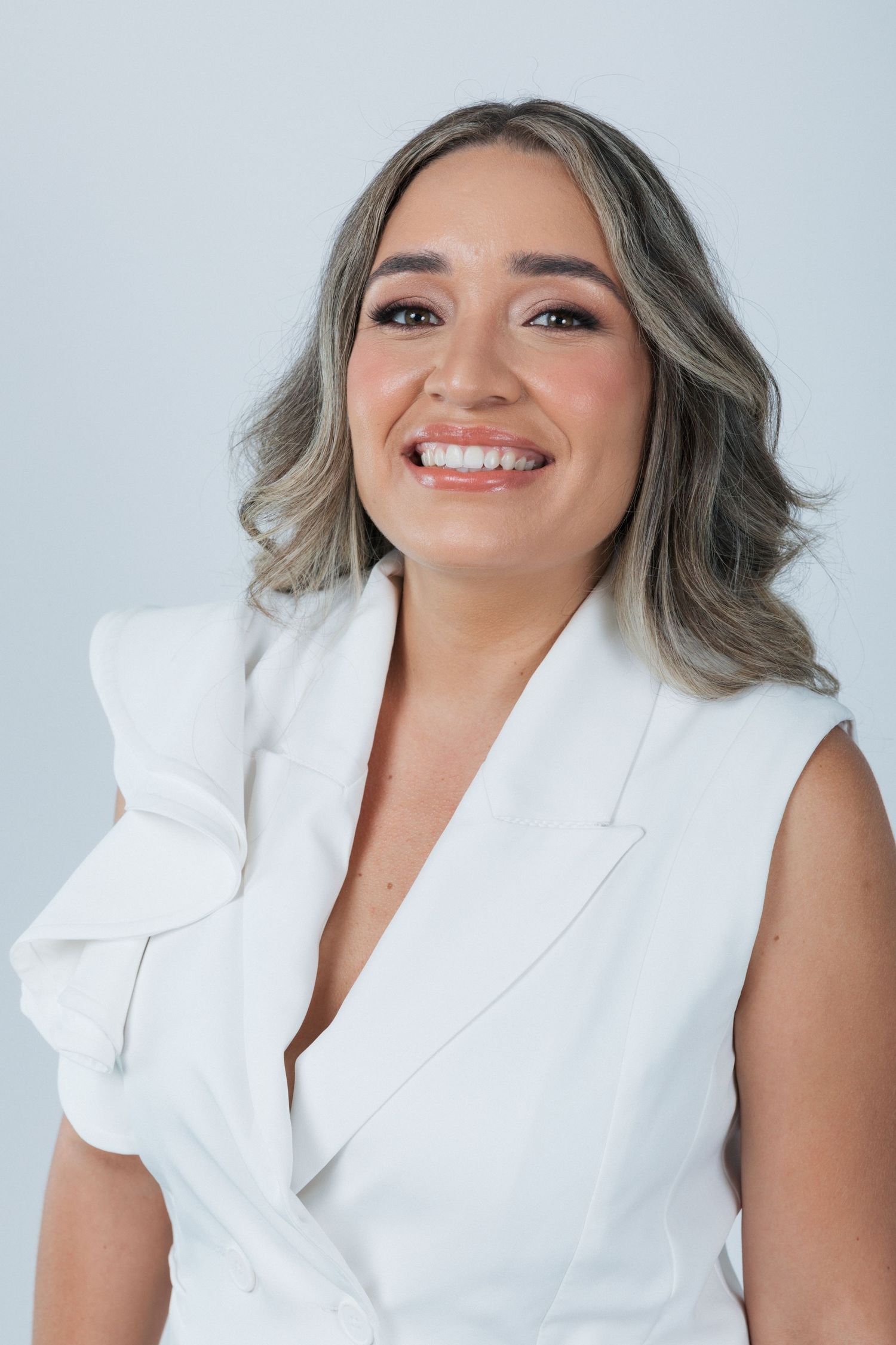 Portrait of a person in a sleeveless white blazer smiling warmly against a neutral backdrop.